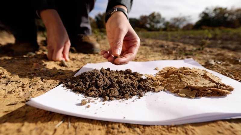 White printer paper is lying on the dirt in an agricultural Two piles of dirt on top of white paper. The one on the left is a rich brown and has a crumbly texture, the one on the right is a light tan and has large flakes of soil crust visible.