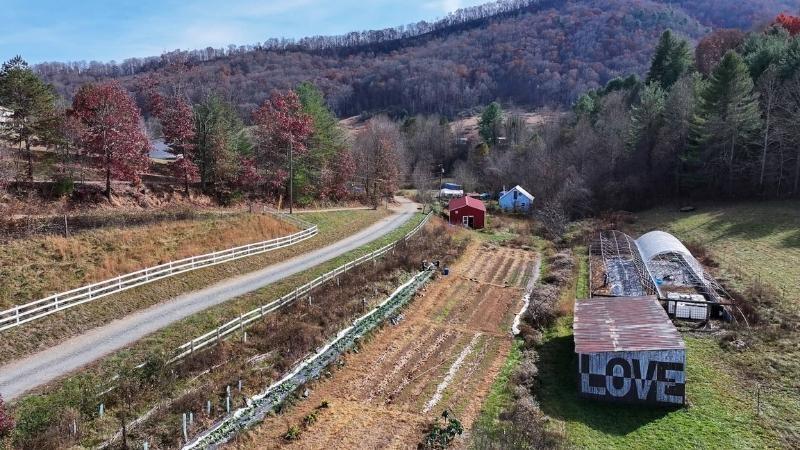An aerial view of a small farm with planted and unplanted beds, a red shed, plastic-covered high tunnels and a wooden barn with the word LOVE painted on the side in black and white. Mountains rise in the distance, and a road runs adjacent to the farm.