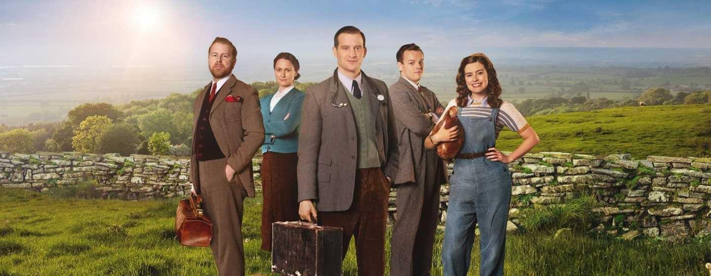 The cast of All Creatures Great & Small poses with the English countryside in the background, a golden retriever at their feet and a small lamb in the foreground.