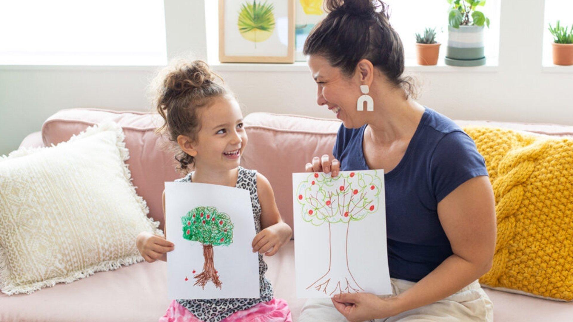 A little girl and a woman sitting on a couch. They're both holding up drawings of an apple tree.