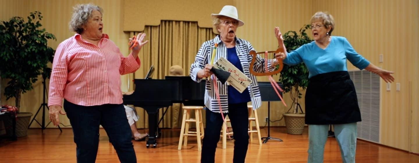 Three women rehearse a musical number in front of another woman playing piano