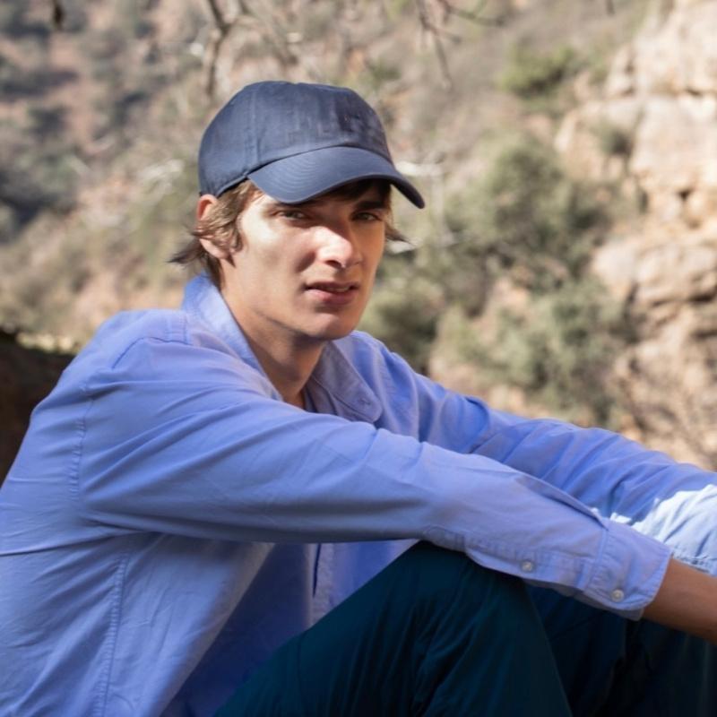 A headshot of Max Feliu in a blue-collared shirt and black baseball cap.