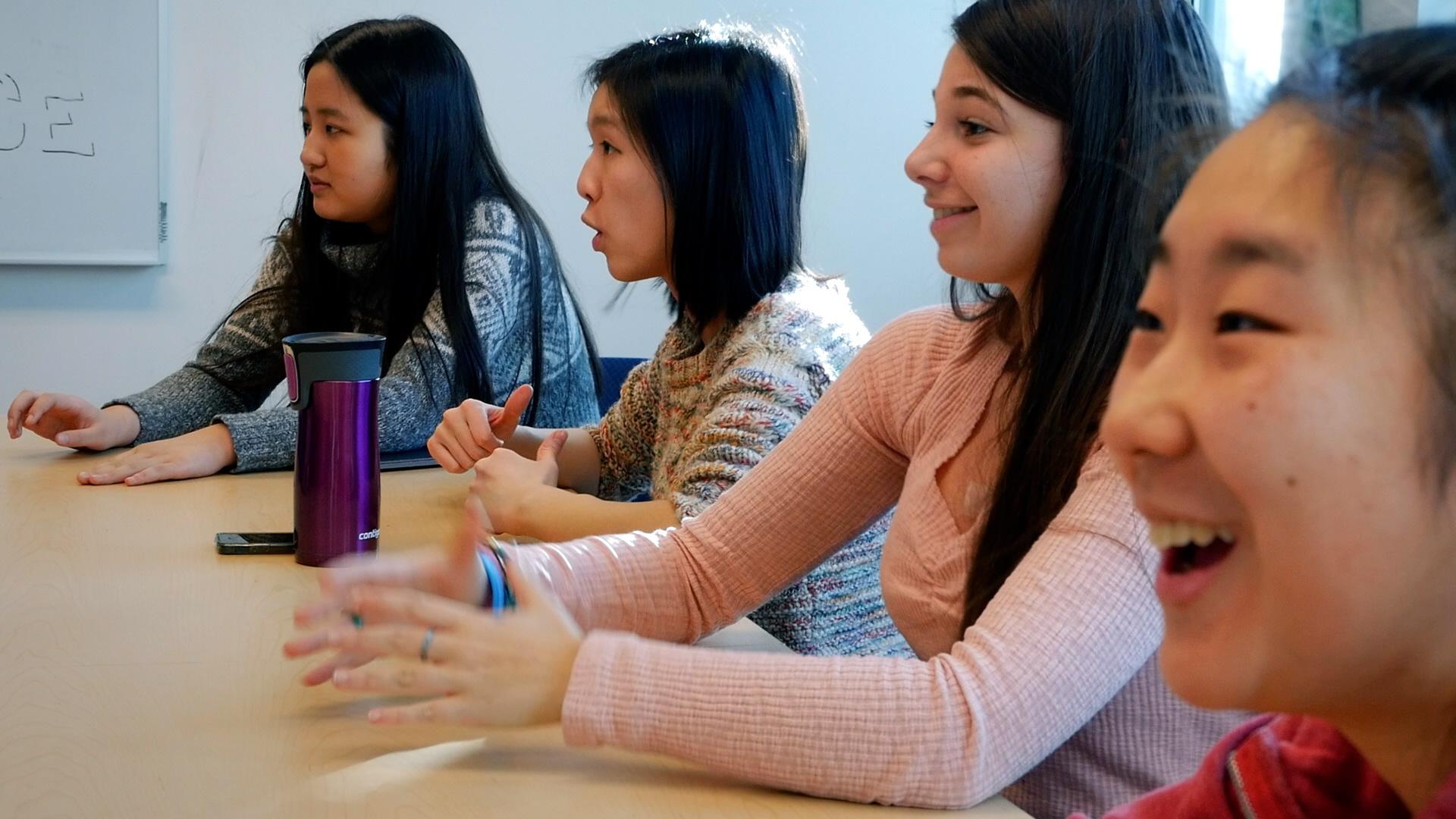 A row of high school girls sit at a table practicing for competitive debate. 