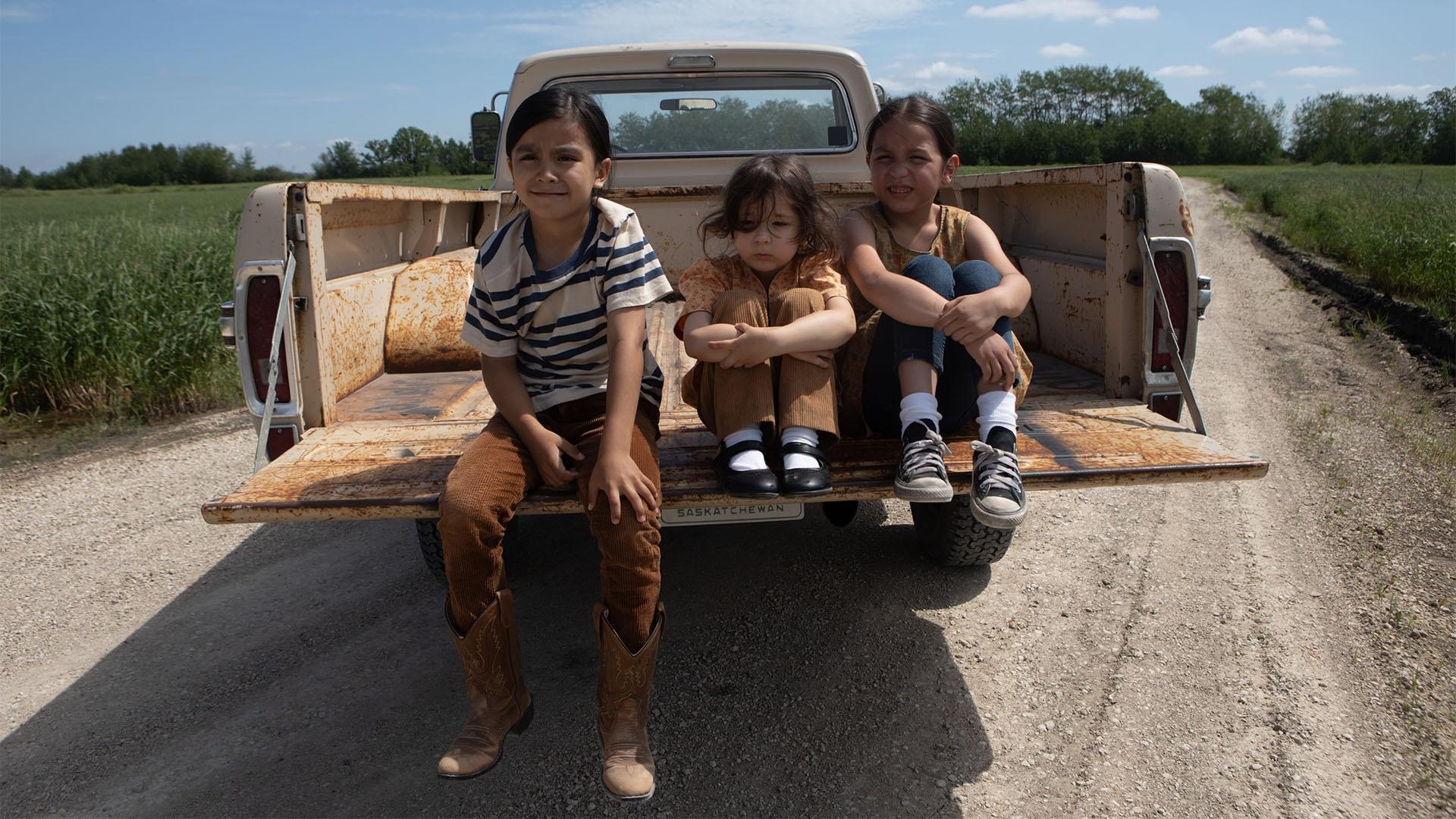 Young Niizh, young Dora, and young Bezhig sit on the back of a truck.