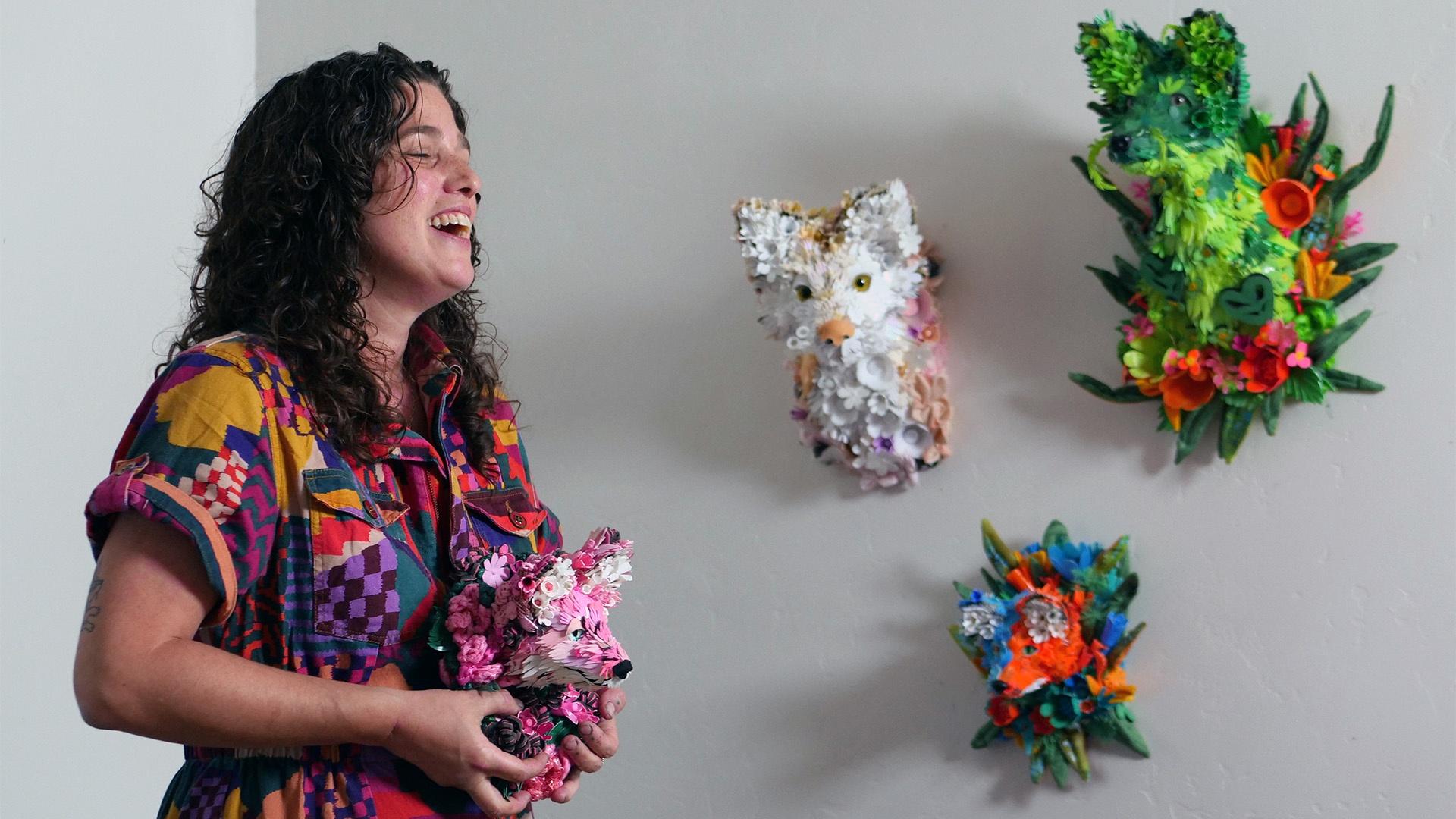 Artist Calder Kamin wearing colorful jumpsuit holds one of her sculptures made of recycled plastic