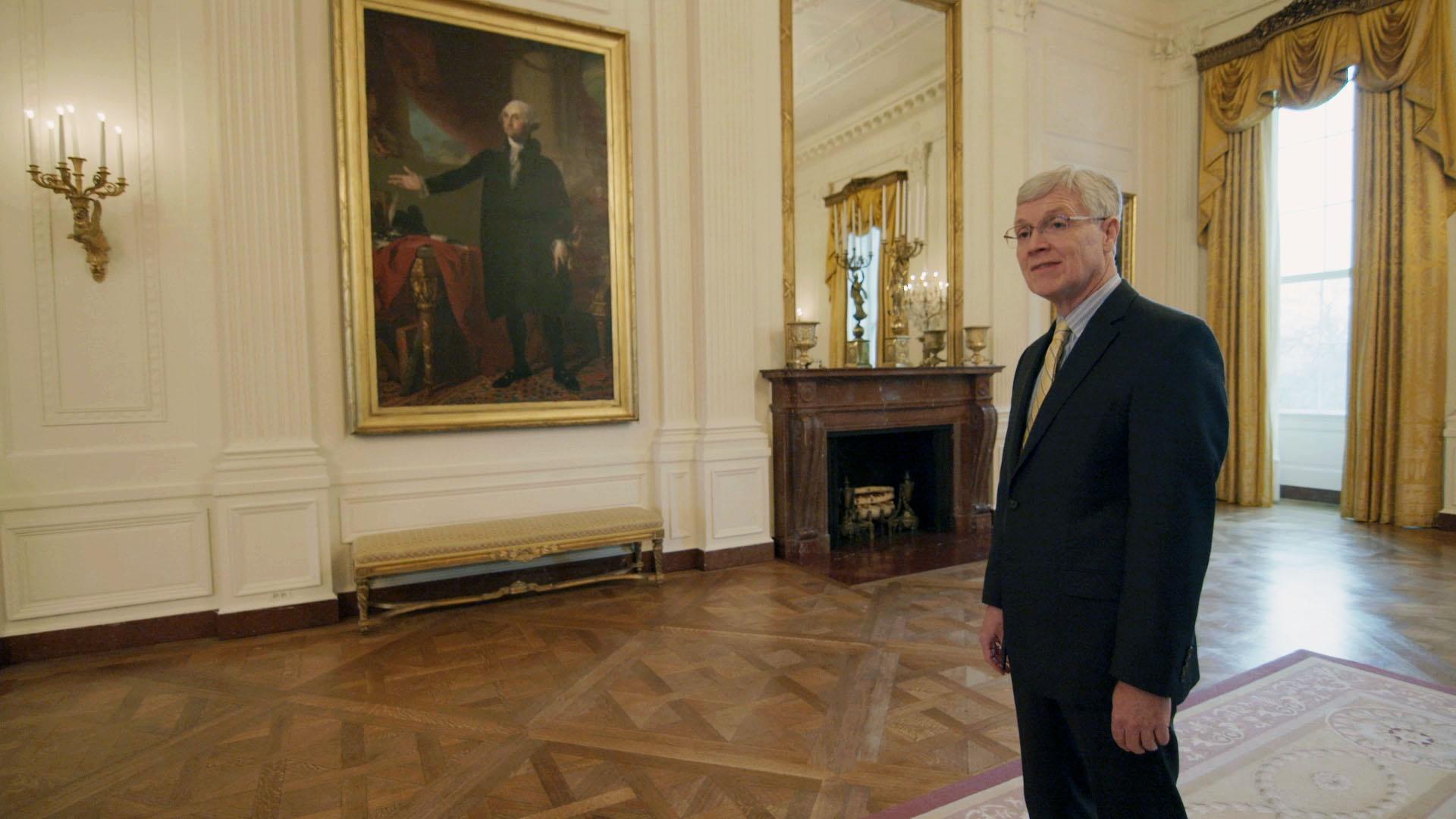 White House Curator Bill Allman stands in East Room of White House with George Washington portrait.
