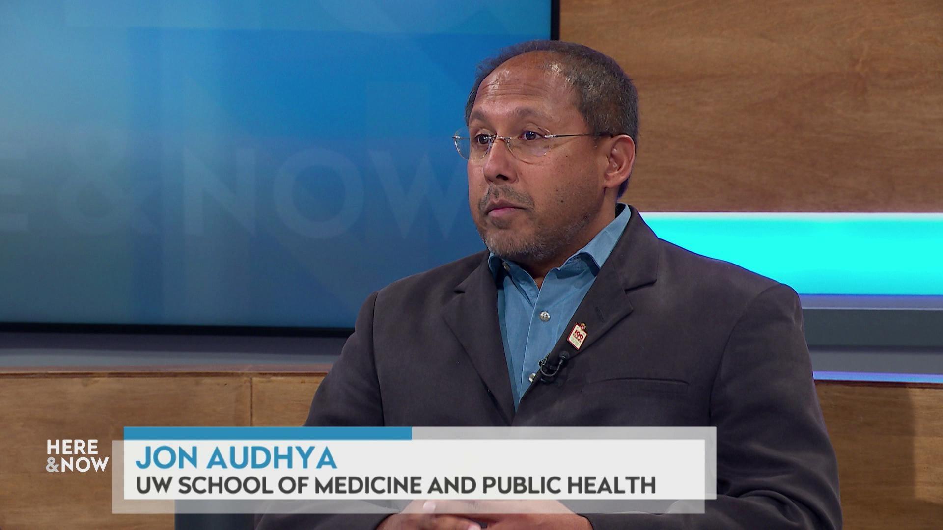 A still image shows Jon Audhya seated at the 'Here & Now' set featuring wood paneling, with a graphic at bottom reading 'Jon Audhya' and 'UW School of Medicine and Public Health.'