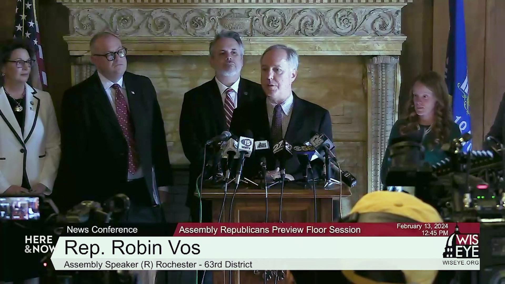 A still image shows Robin Vos standing behind a podium and microphones addressing a room of people with people standing alongside him on either side.