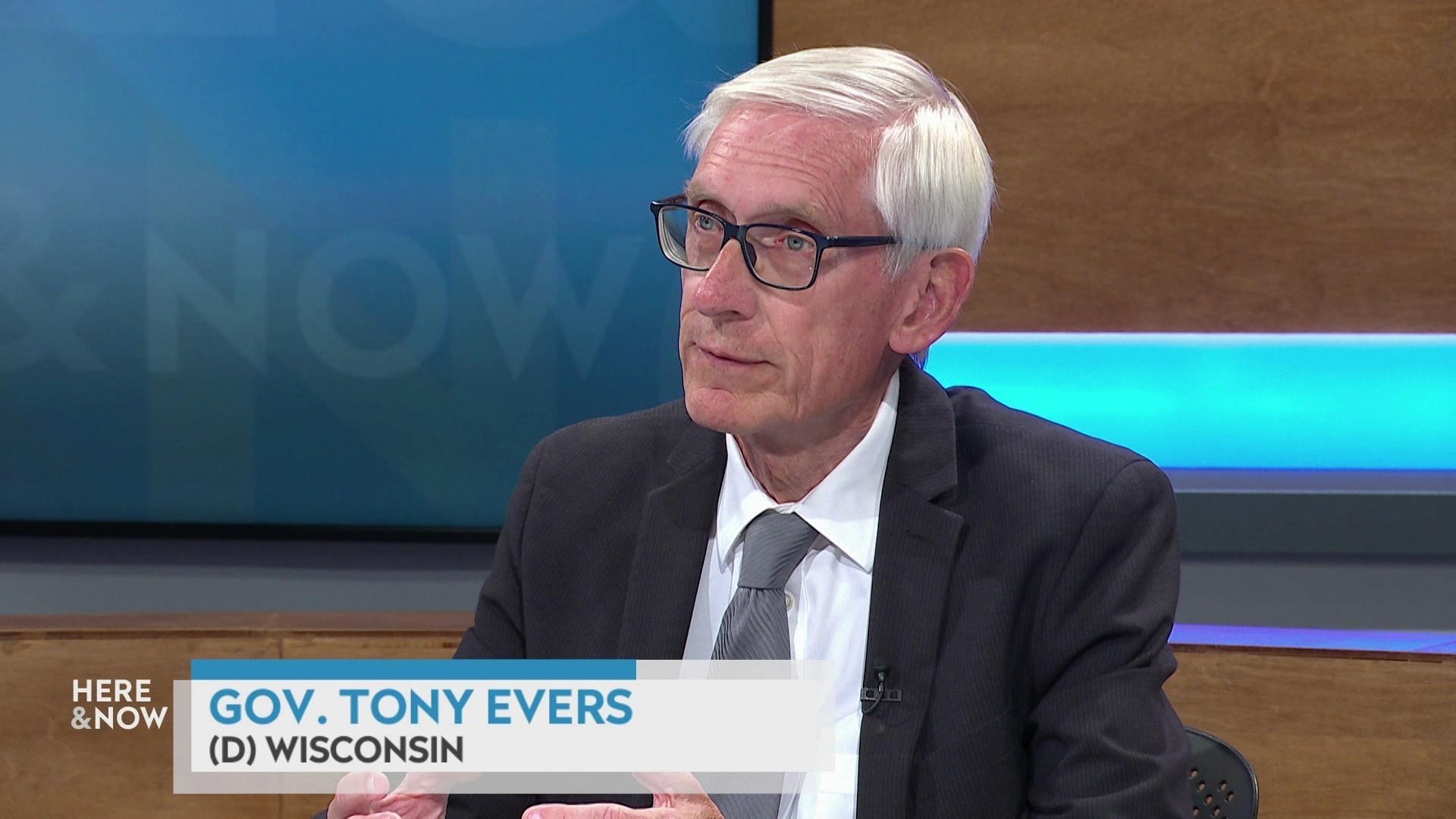 A still image shows Tony Evers seated at the 'Here & Now' set featuring wood paneling, with a graphic at bottom reading 'Gov. Tony Evers' and '(D) Wisconsin.'