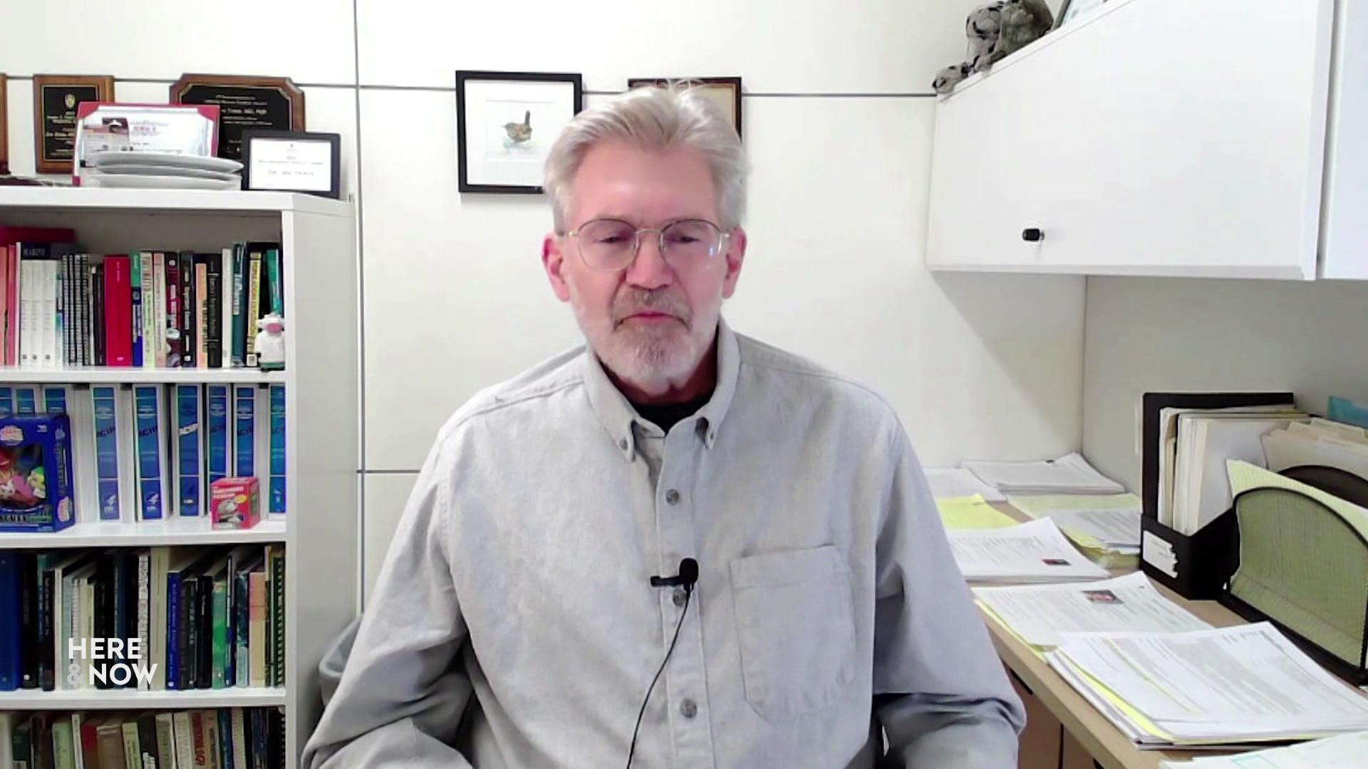 A still image from a video shows Johnathan Temte seated in front of a desk with piles of papers and a bookshelf lined with books and other items.