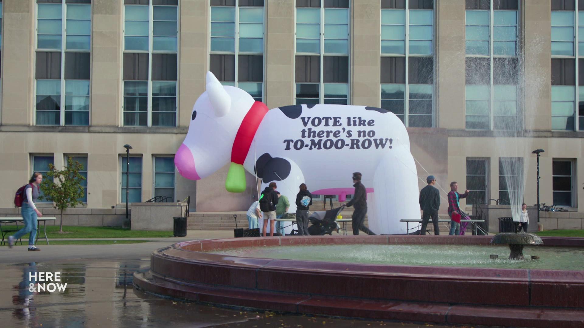 An inflatable cow with the words 'Vote like there's no to-moo-row!' sits on a patch of grass with a building with glass windows in the background, people walking around and a fountain in the foreground.