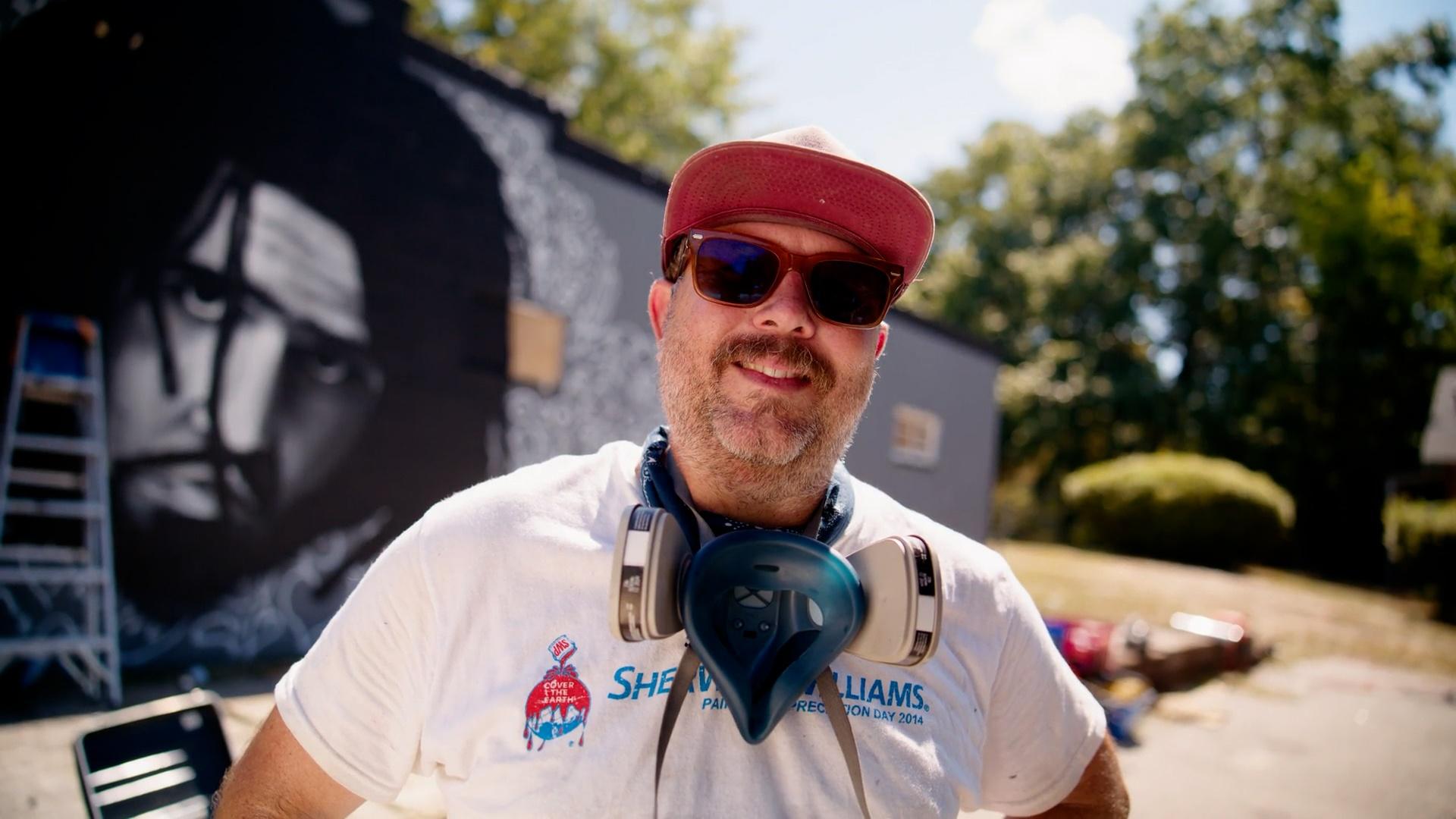 A muralist in front of a mural of J. Cole.