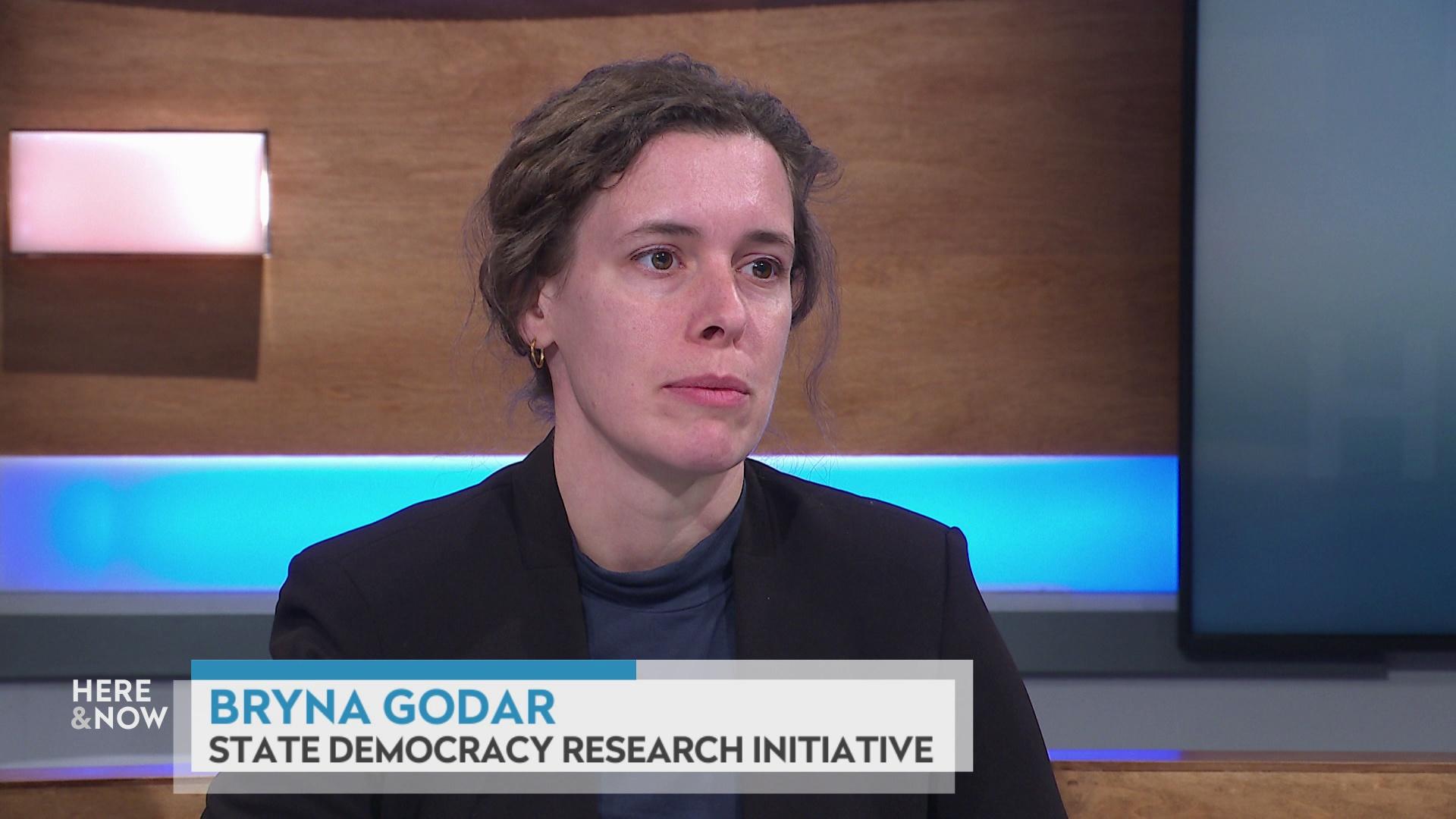 A still image shows Bryna Godar seated at the 'Here & Now' set featuring wood paneling, with a graphic at bottom reading 'Bryna Godar' and 'State Democracy Research Initiative.'