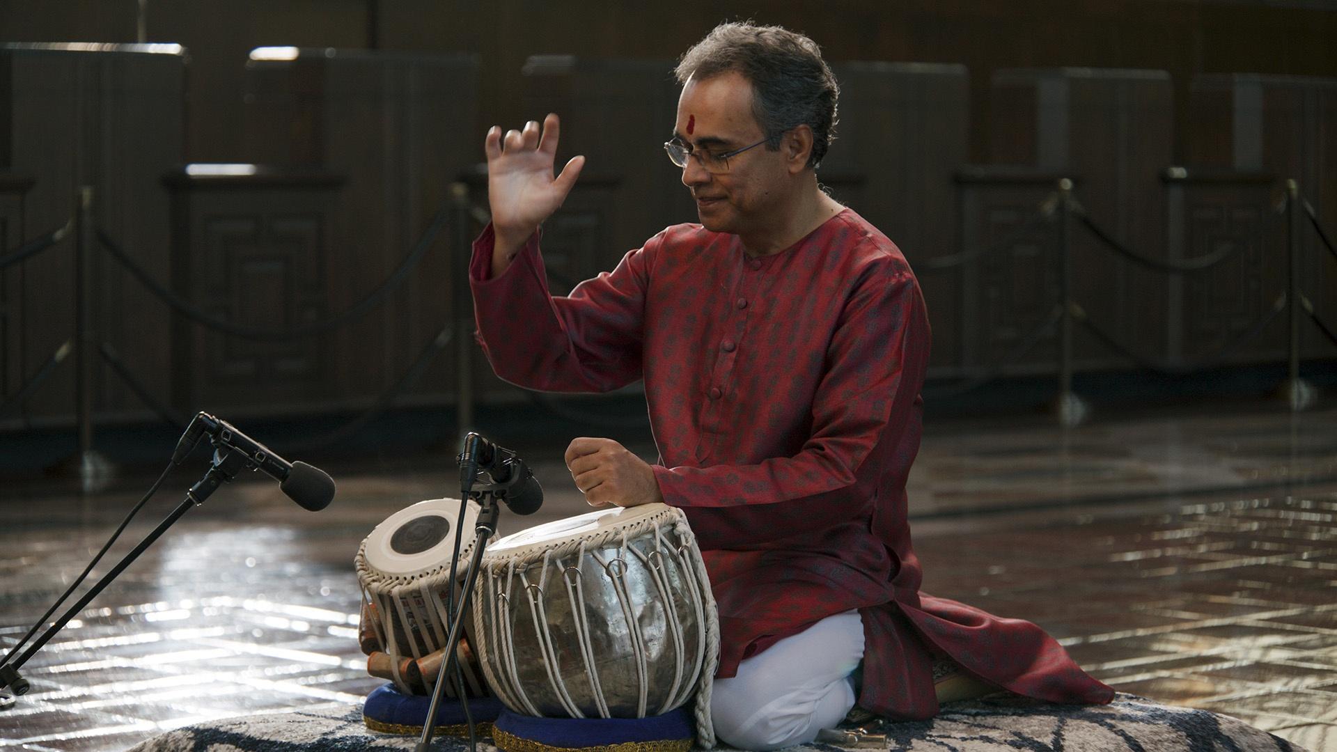 Sandeep Das playing bongos in front of microphones while kneeling on a gymnasium floor.