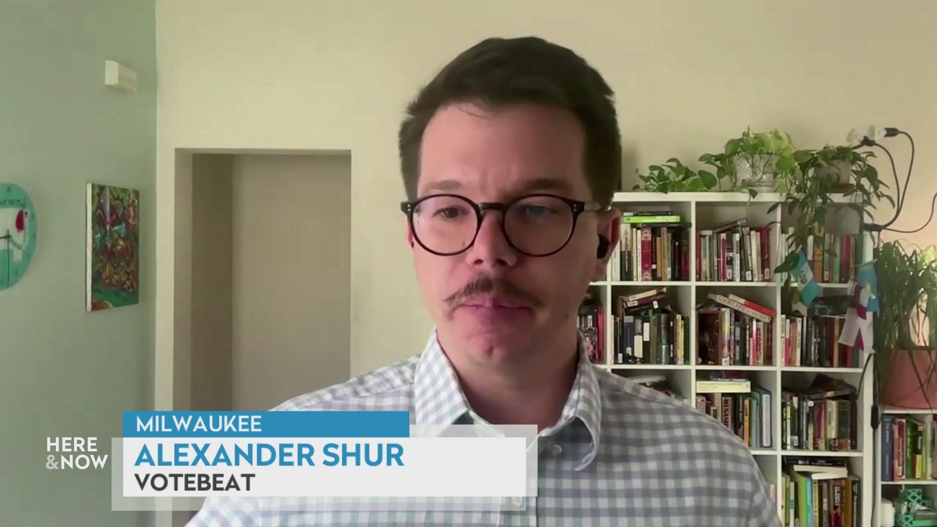 A still image shows Alexander Shur standing in front of a bookcase with an ivy plant on top.