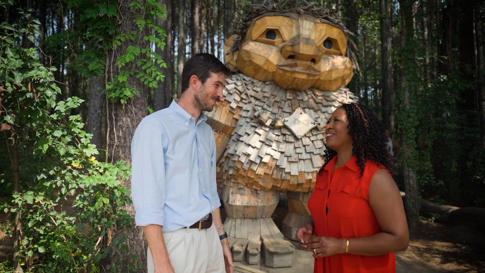 Host of NC Weekend Deborah Holt Noel next to a tall white man, in front of one of the wooden trolls located at Dix Park in Raleigh.