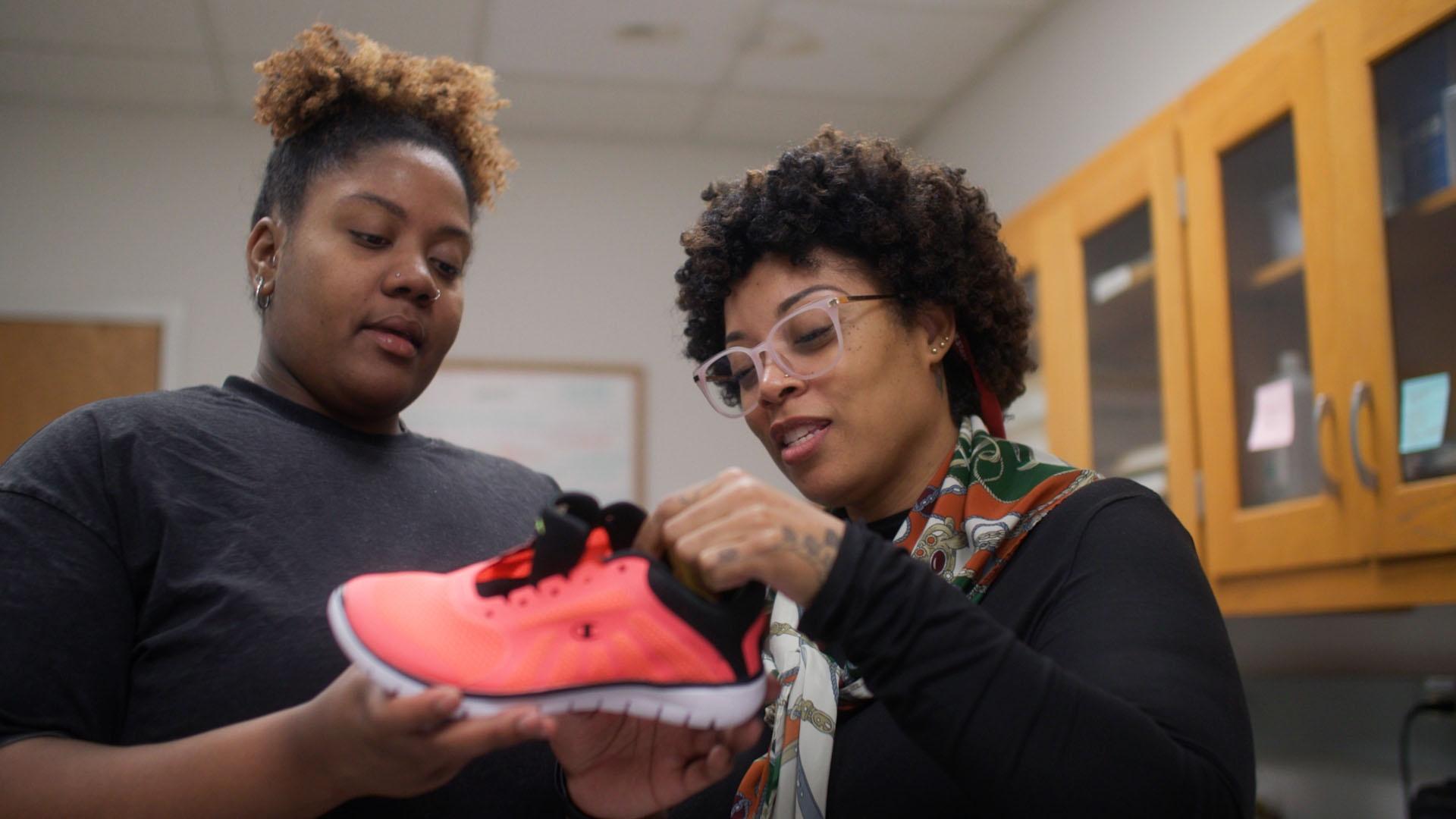 Two Black women standing together in a lab holding up a bright pink exercise sneaker.