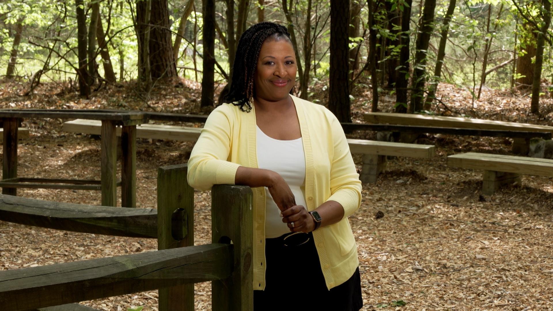 Deborah Holt Noel, host of NC Weekend, leans up against a wooden fence in the forest.