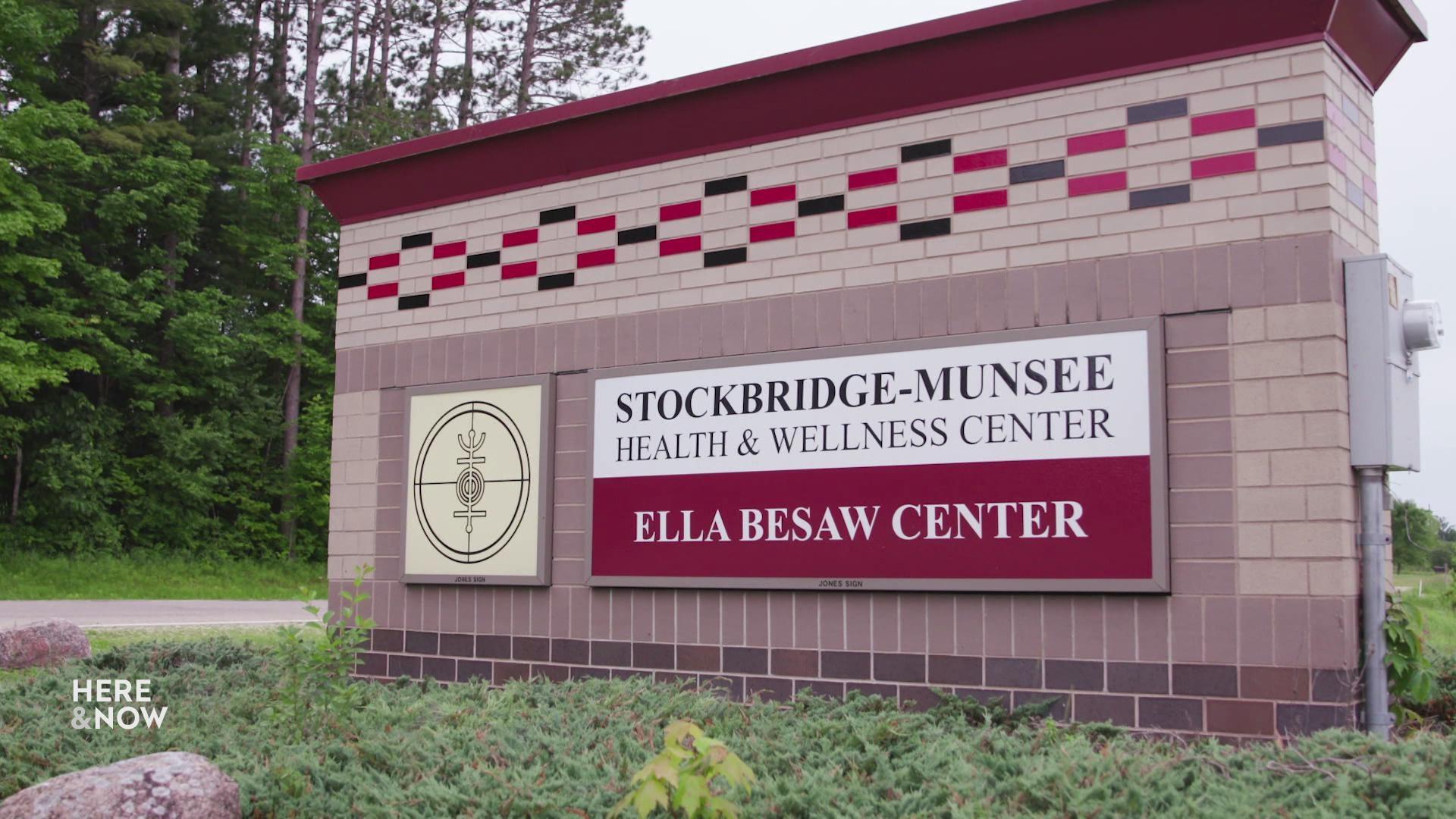 A still image shows a brick road sign with red and black patterns in a bushy area, the sign reads 'Stockbridge-Munsee Health & Wellness Center' and 'Ella Besaw Center.'