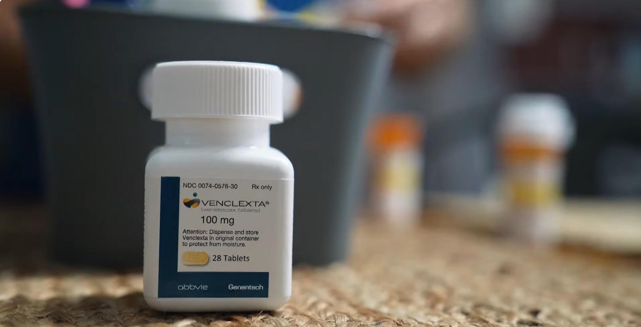 A plastic medication bottle with a label reading Venclexta®, 100 mg and 28 tablets sits on a woven fiber surface, with two out-of-focus medication vials in the background.