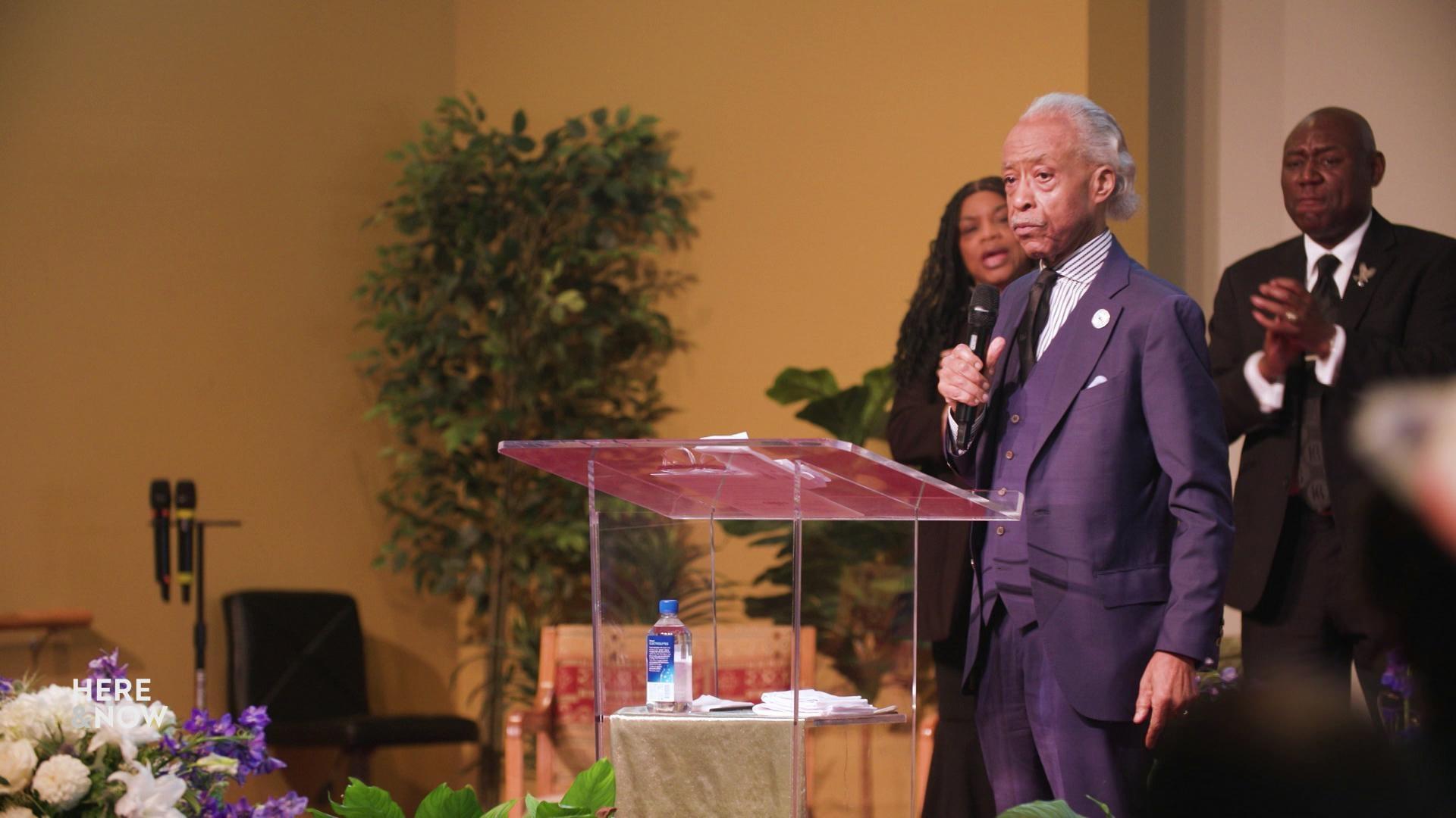 A still image shows Rev. Al Sharpton giving a eulogy at a podium at Corey Ruiz's funeral, with Ben Crump behind him applauding.