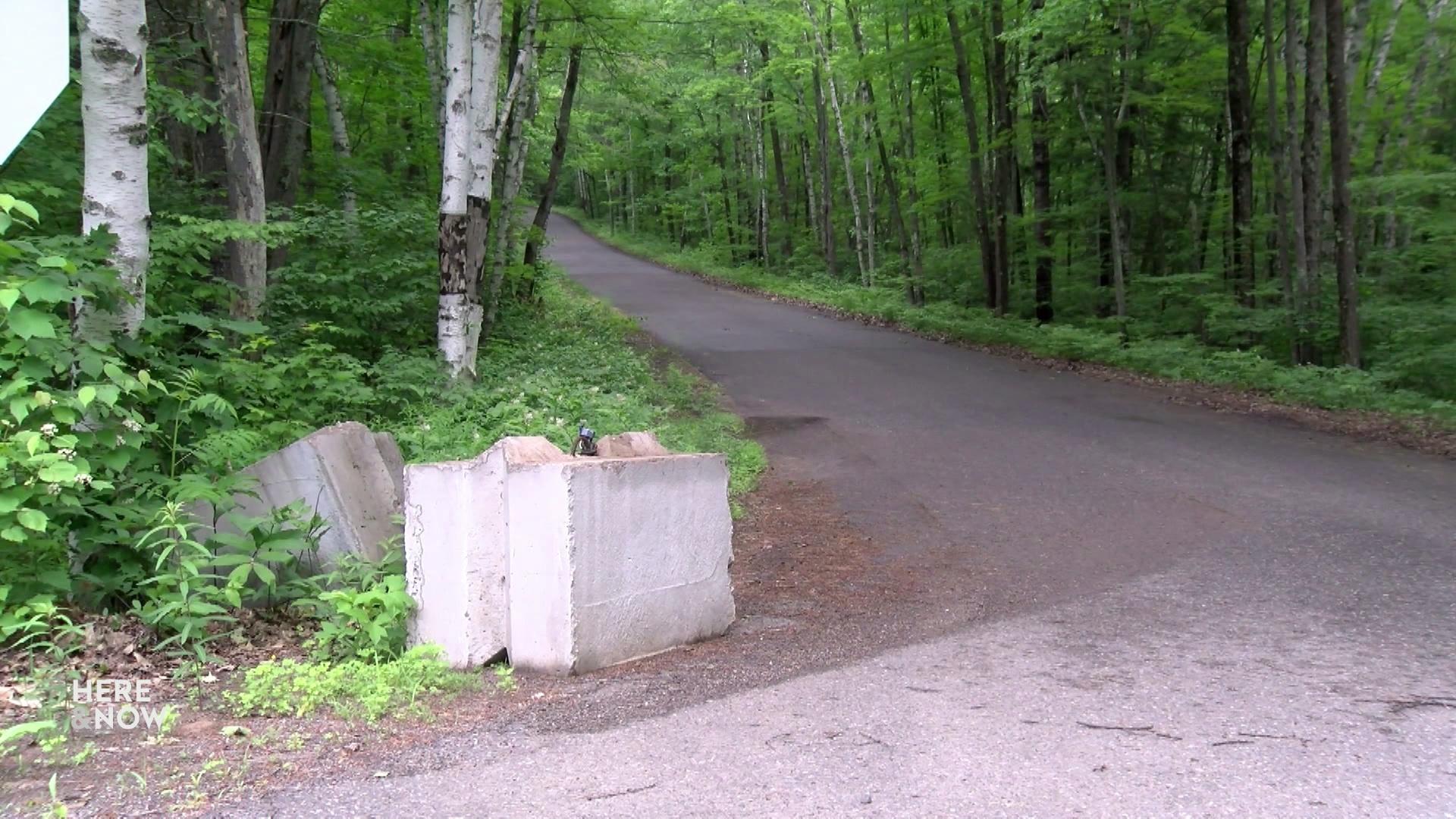 A still image shows concrete blocks on the side of a road running through a wooded area with trees and dense undergrowth.