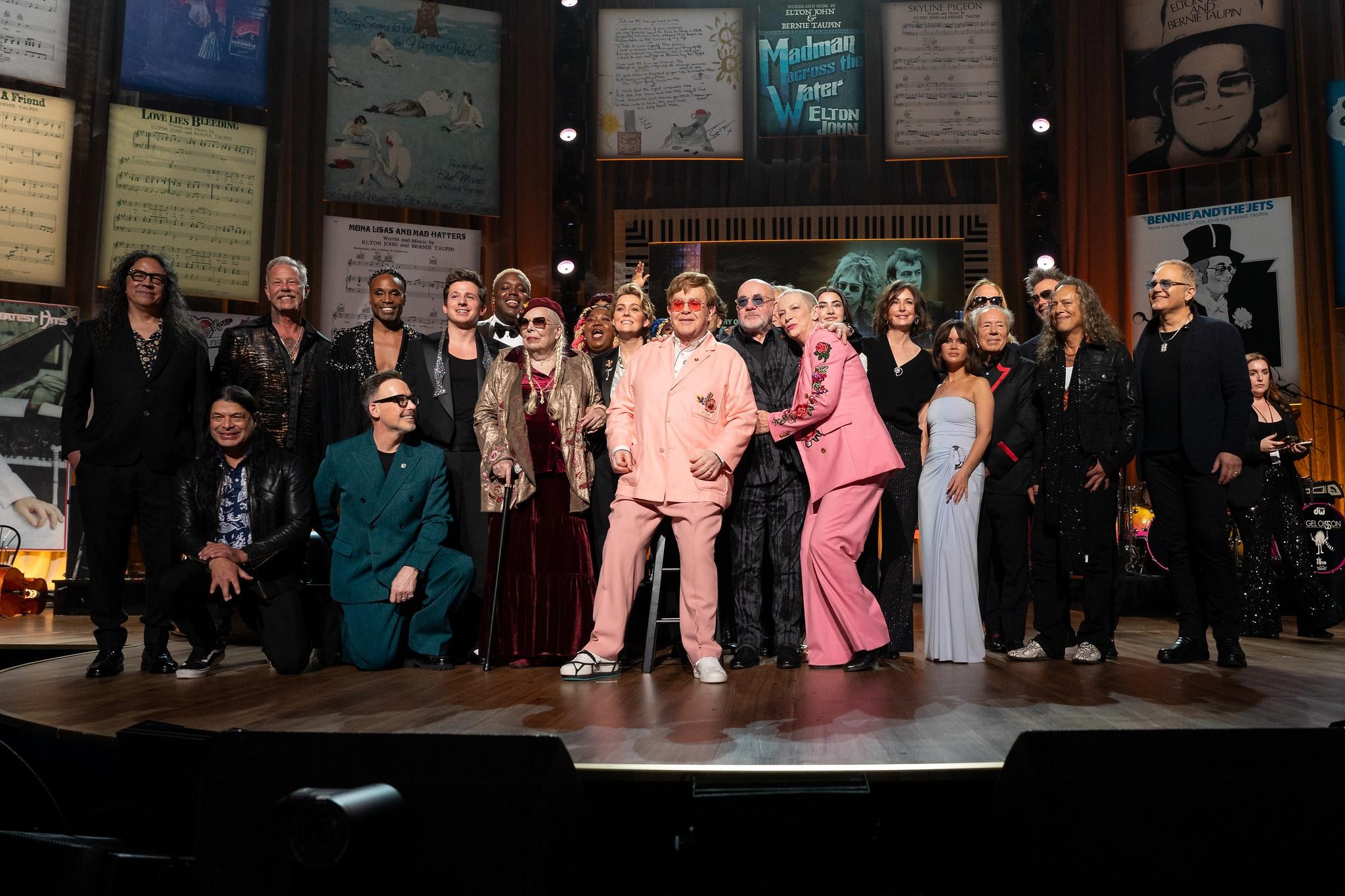 Elton John in pink suit and Bernie Taupin surrounded by cast of other entertainers
