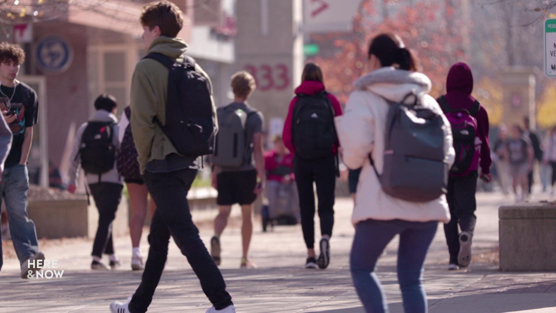 A still image shows various students with backpacks walking on a paved street outside.