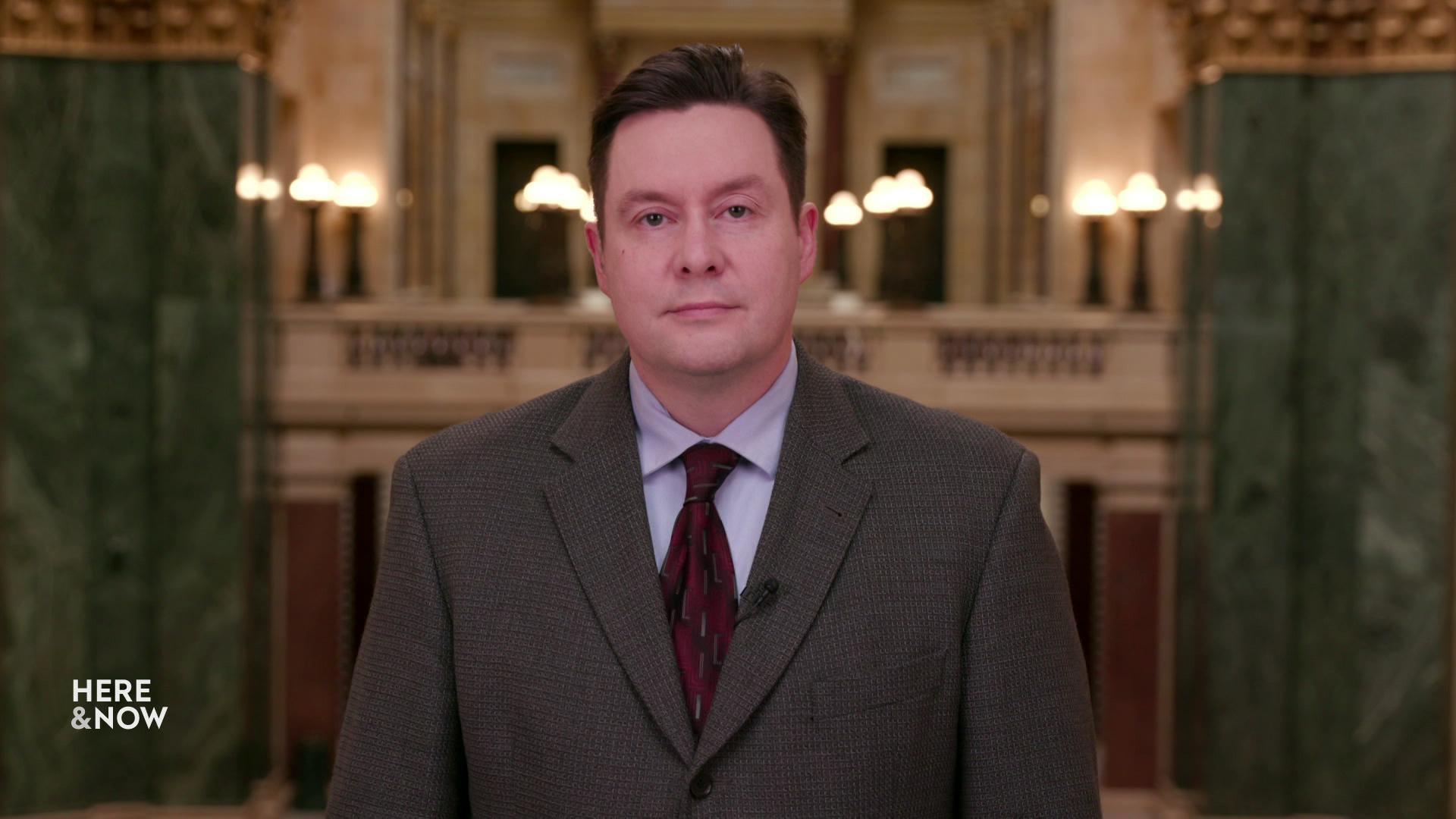 A still image shows Zac Schultz standing in the state Capitol building with two large green stone pillars on either side. A still image shows Zac Schultz standing in the state Capitol building with two large green stone pillars on either side.