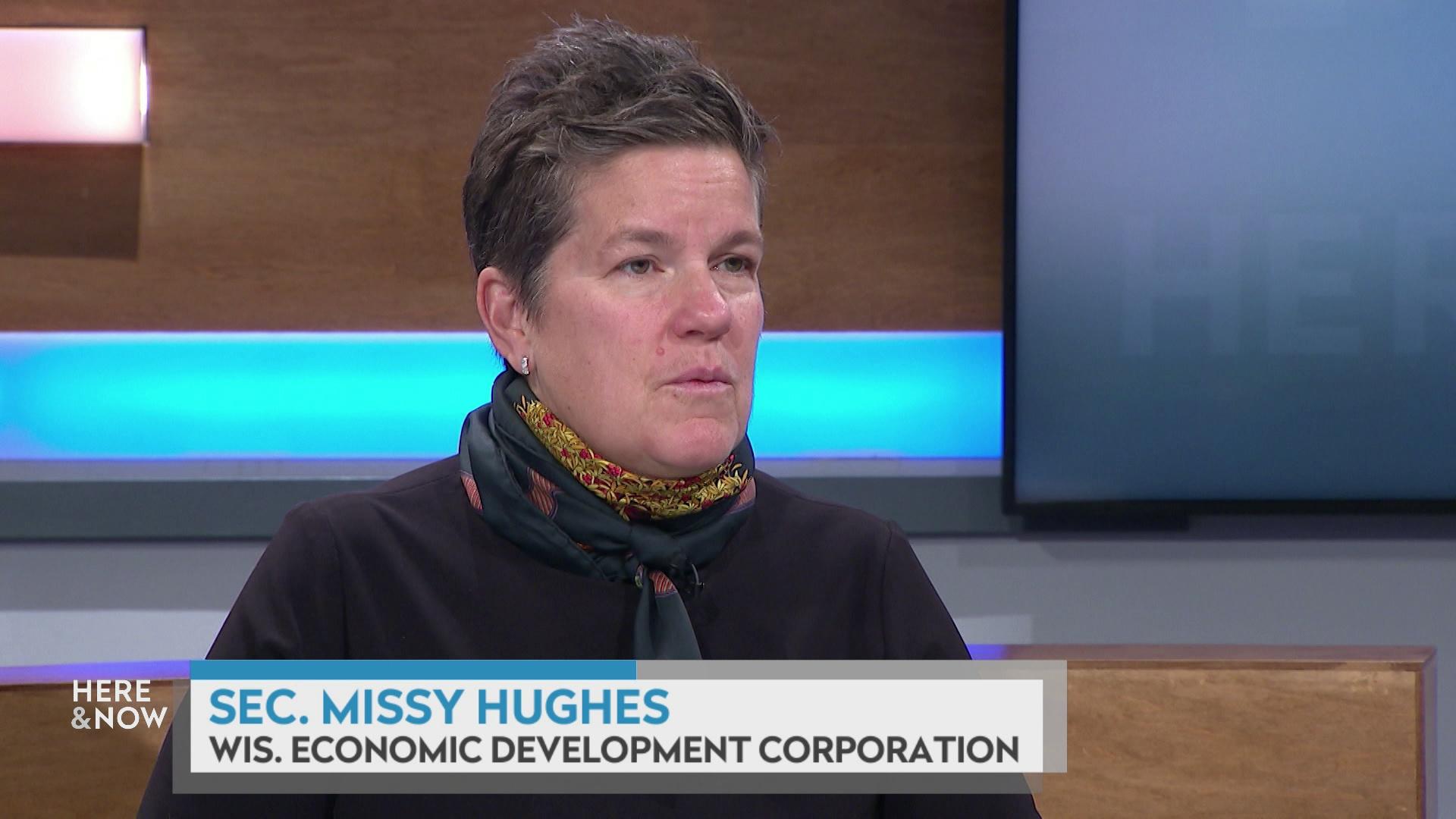 A still image shows Missy Hughes seated at the 'Here & Now' set featuring wood paneling, with a graphic at bottom reading 'Missy Hughes' and 'Wis. Economic Development Corporation.'