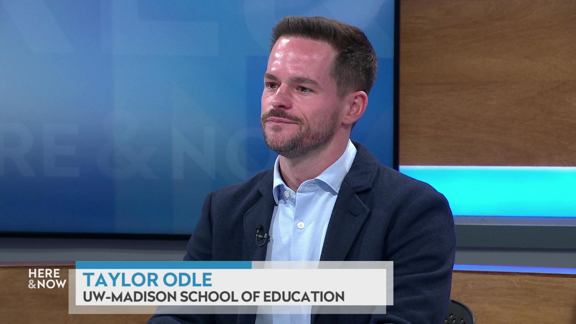 A still image shows Taylor Odle seated at the 'Here & Now' set featuring wood paneling, with a graphic at bottom reading 'Taylor Odle' and 'UW-Madison School of Education' A still image shows Taylor Odle seated at the 'Here & Now' set featuring wood paneling, with a graphic at bottom reading 'Taylor Odle' and 'UW-Madison School of Education'