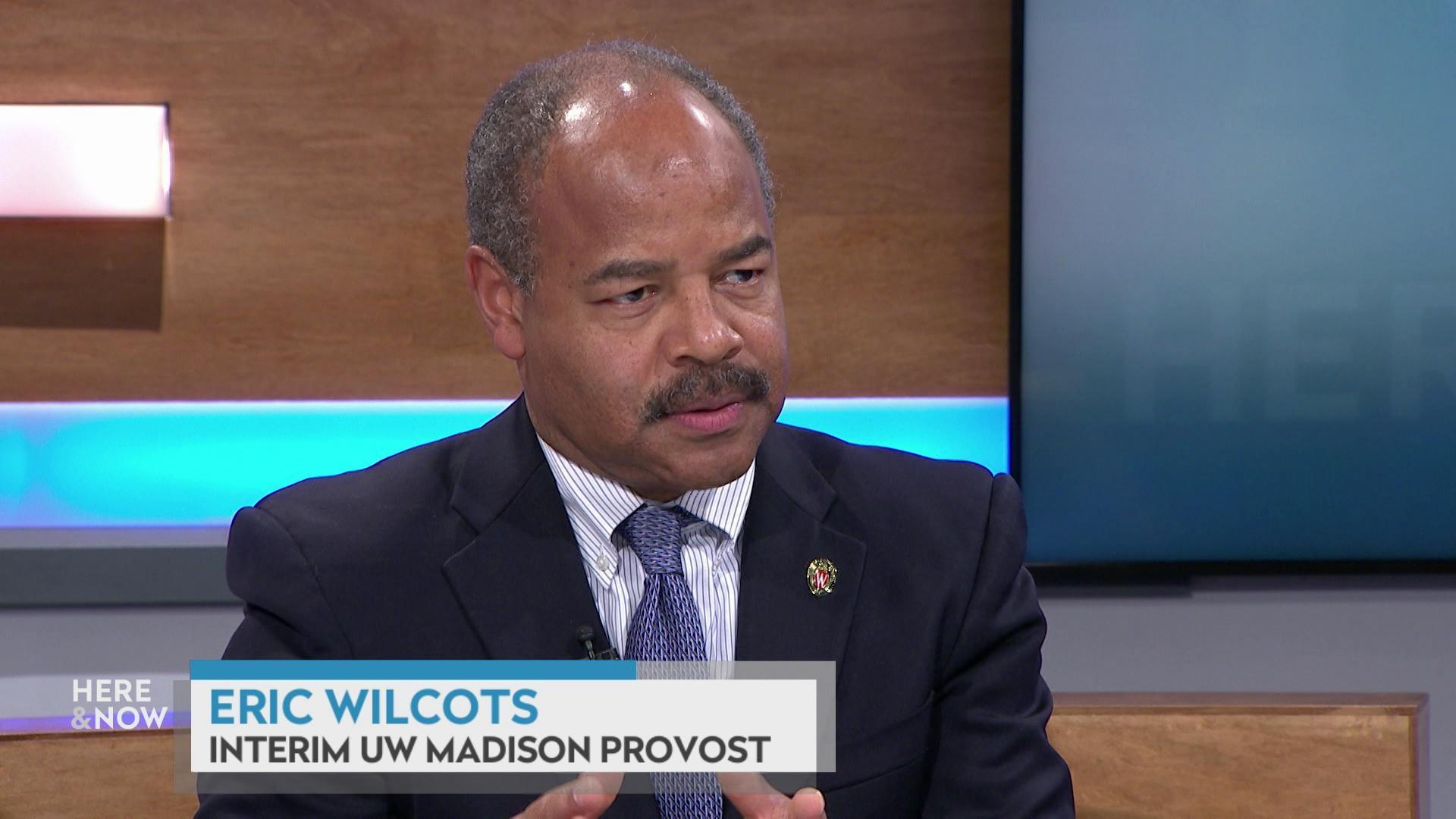 A still image from a video shows Eric Wilcots seated at the 'Here & Now' set featuring wood paneling, with a graphic at bottom reading 'Eric Wilcots' and 'Interim UW Madison Provost.'