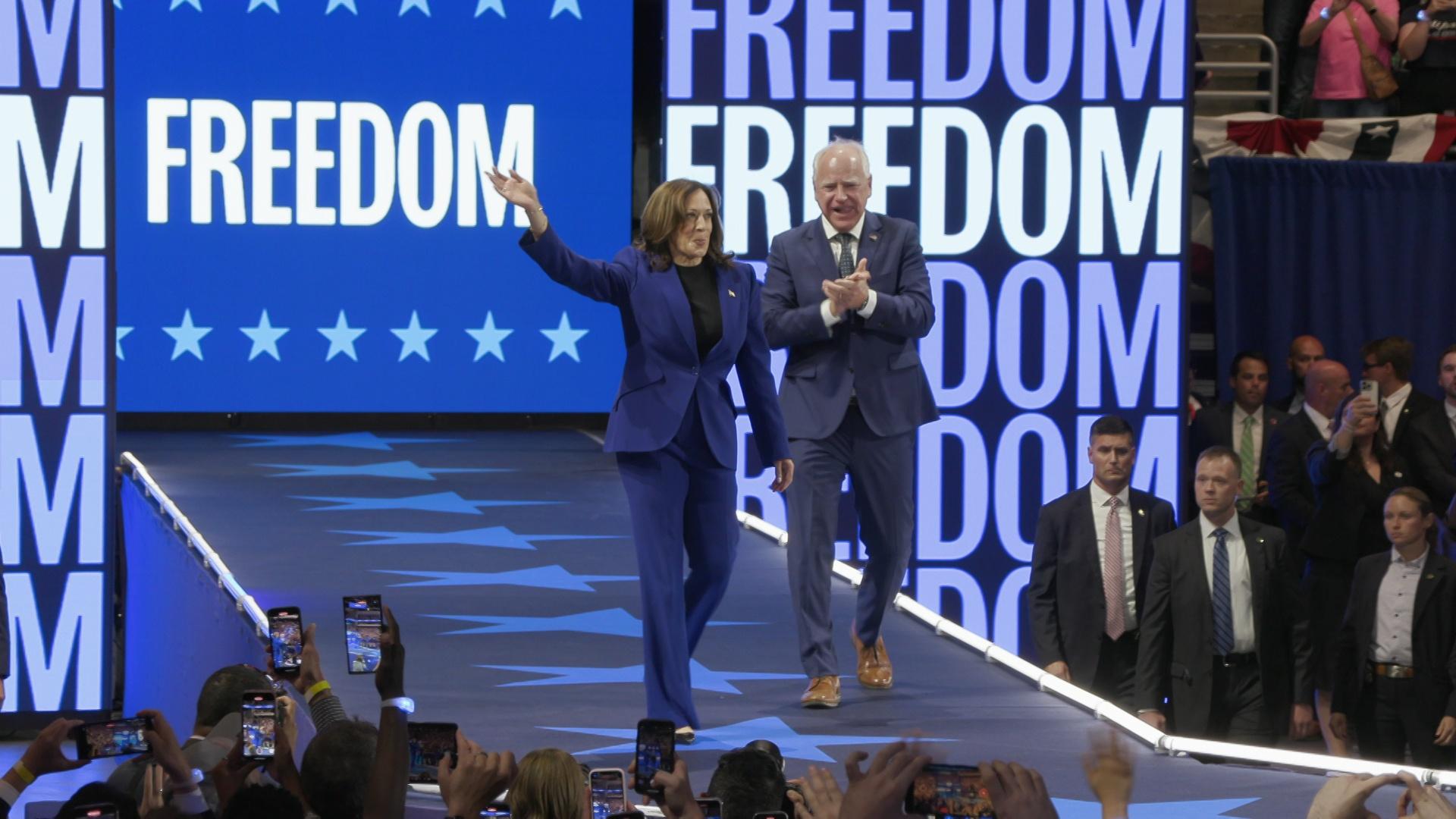 Kamala Harris and Tim Walz walk and gesture indoors on a runway with screens reading 'Freedom' and people standing below.