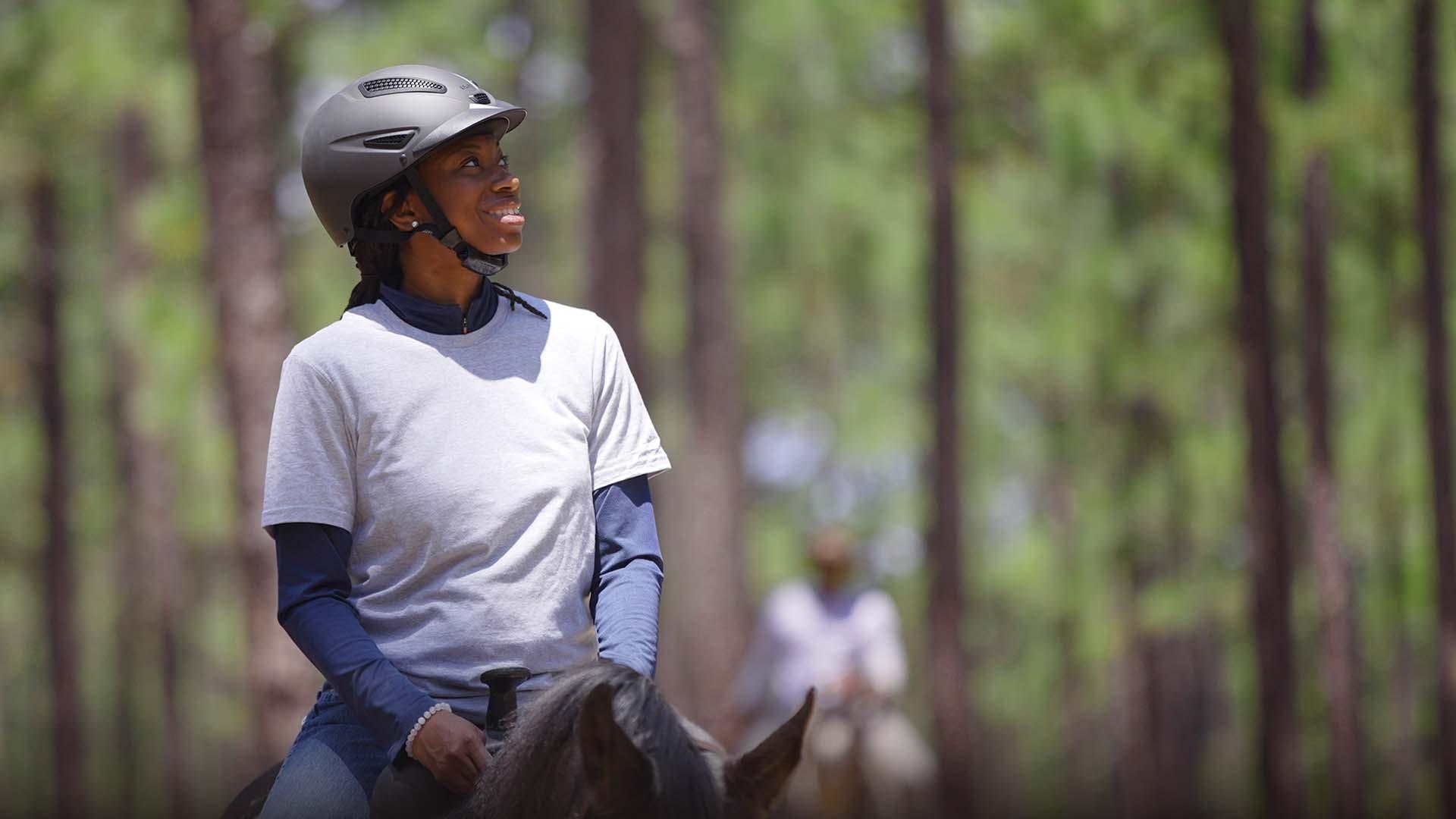 Caitlin Gooch with a helmet on riding on a horse through the woods.