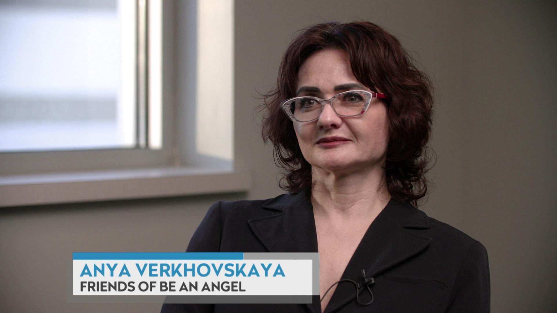 A still image from a video shows Anya Verkhovskaya sitting in a room with a window behind her right shoulder, with a graphic at bottom reading Anya Verkhovskaya and Friends of Be an Angel.