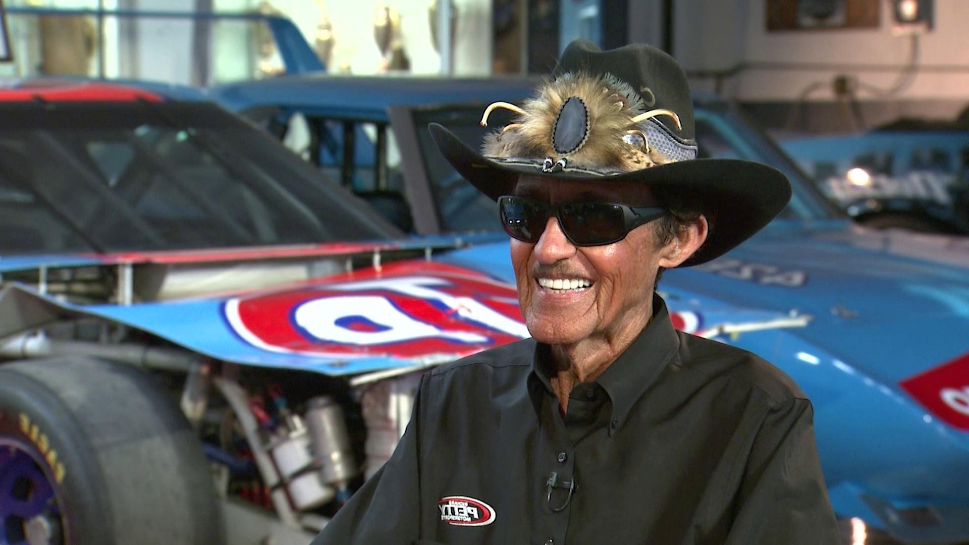 Richard Petty in a cowboy hat smiling with race cars behind him.