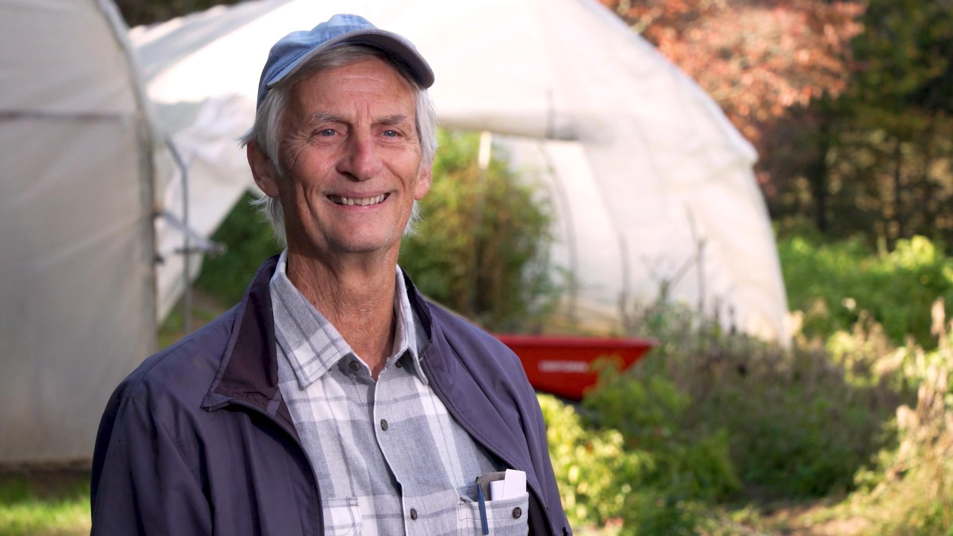 An older white man smiles in front of his farm's greenhouses.