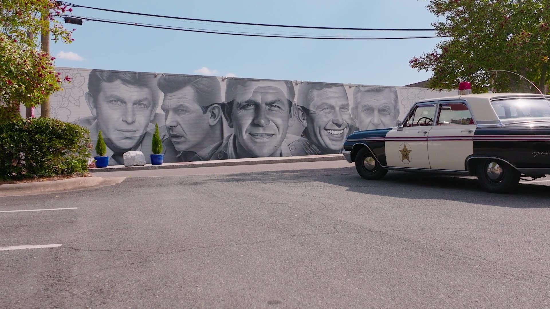 A mural of Andy Griffith with an old police squad car parked in front of it.