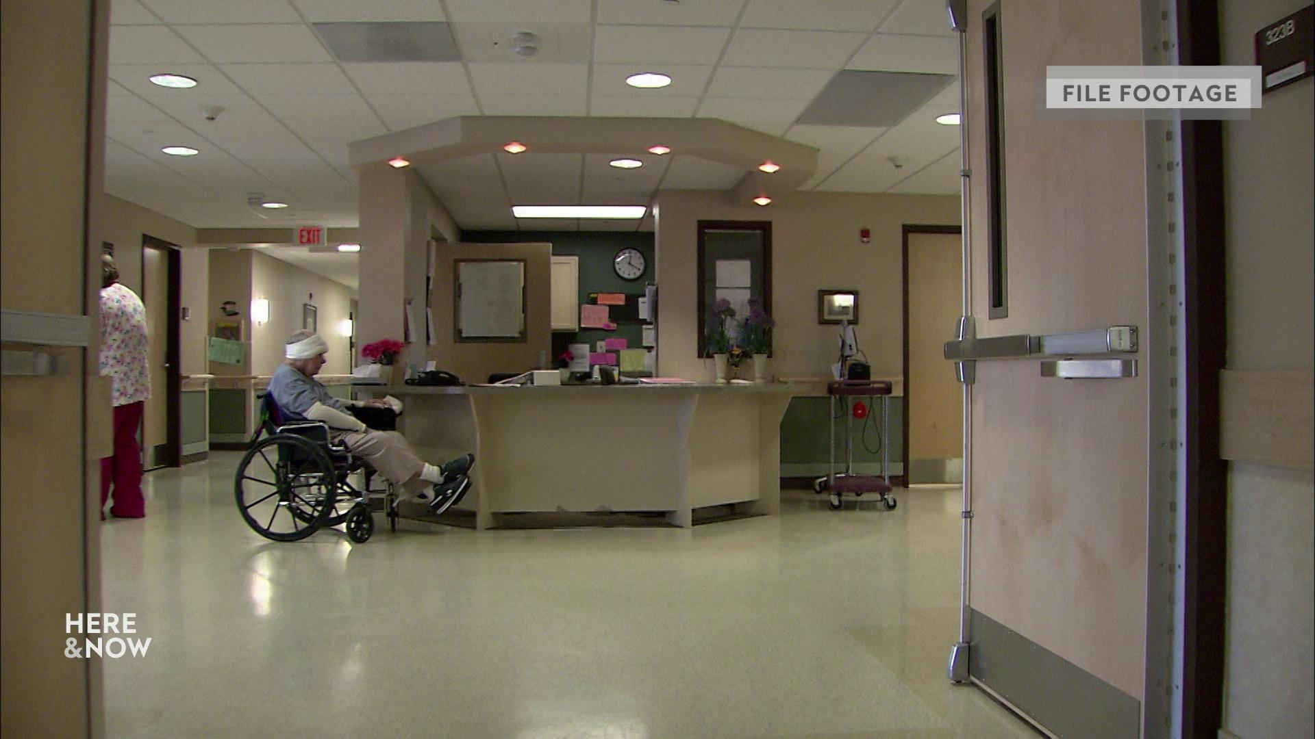 A still image shows a perspective through open doors of a front desk and waiting room in a Wisconsin Veteran Home with a man seated in a wheelchair and a bandaged head.