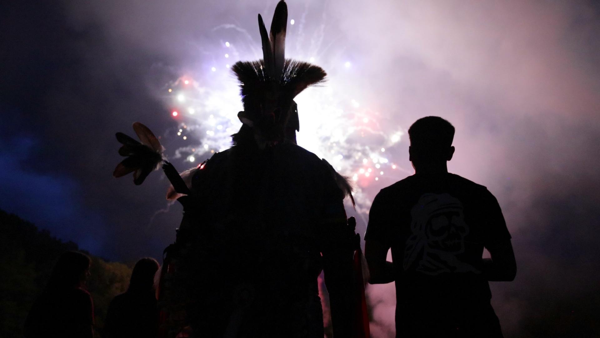 A silhouette of two people in front of fireworks, one is dressed in Native regalia. 