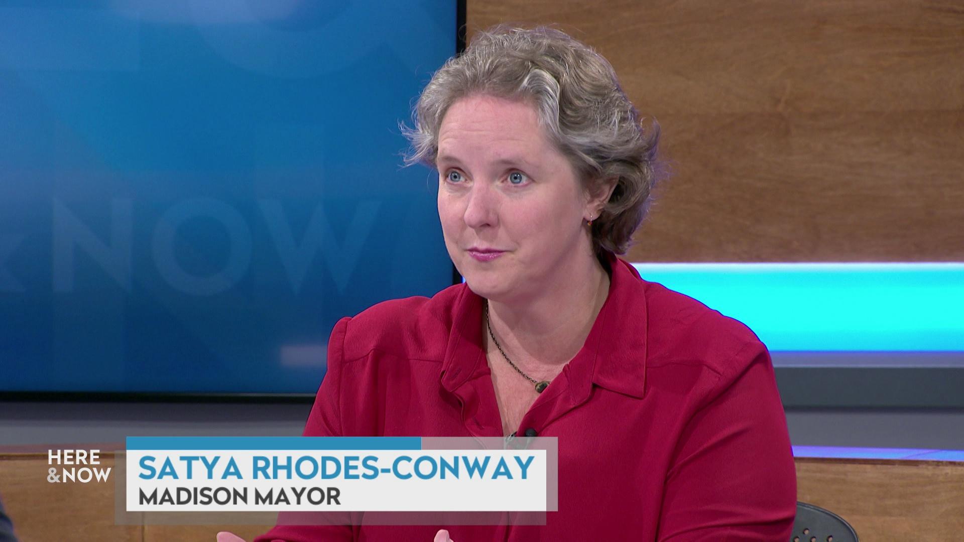 A still image shows Satya Rhodes-Conway seated at the 'Here & Now' set featuring wood paneling, with a graphic at bottom reading 'Satya Rhodes-Conway' and 'Madison Mayor.'