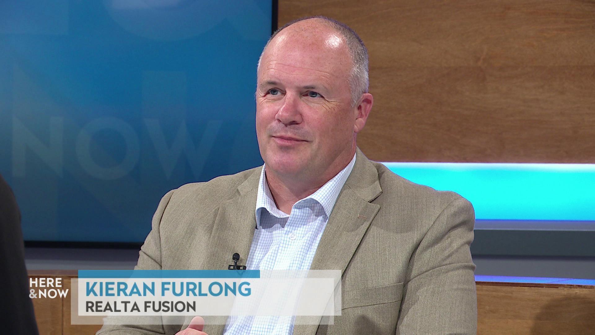 A still image shows Kieran Furlong seated at the 'Here & Now' set featuring wood paneling, with a graphic at bottom reading 'Kieran Furlong' and 'Realta Fusion.' A still image shows Kieran Furlong seated at the 'Here & Now' set featuring wood paneling, with a graphic at bottom reading 'Kieran Furlong' and 'Realta Fusion.'