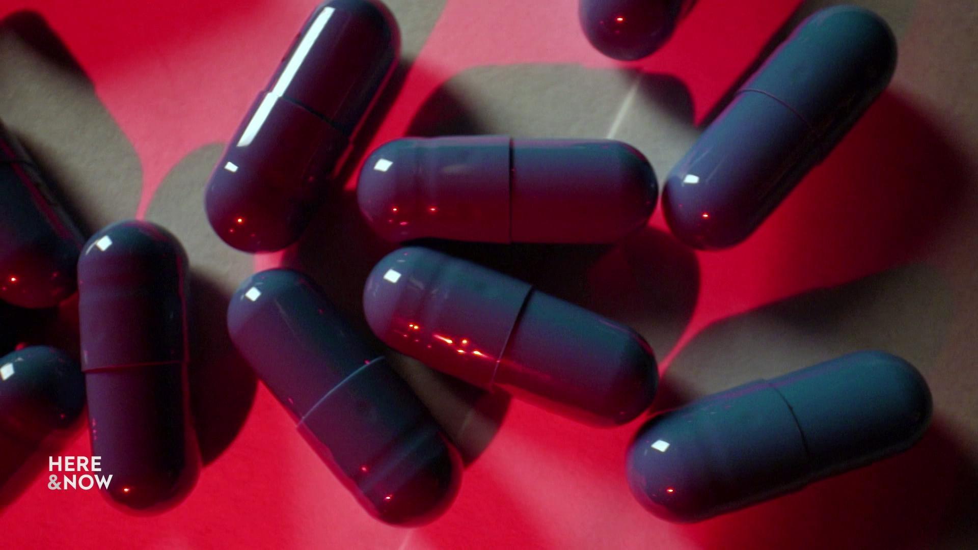 A still image shows an assortment of blue pill capsules underneath a harsh red light.