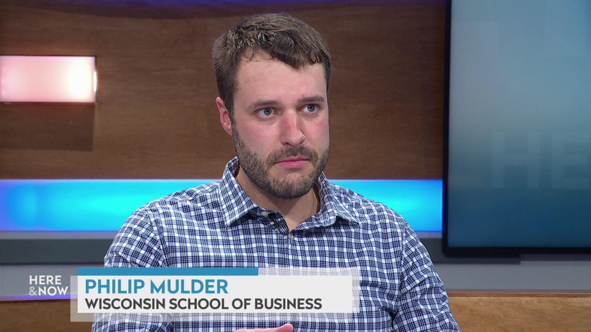 A still image shows Philip Mulder seated at the 'Here & Now' set featuring wood paneling, with a graphic at bottom reading 'Philip Mulder' and 'Wisconsin School of Business.'
