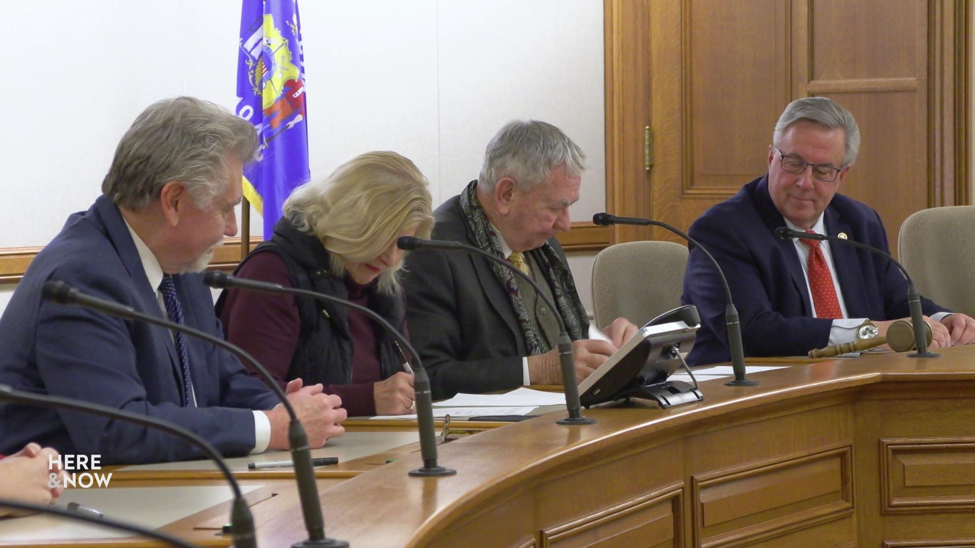 Four people sit behind microphones and a wooden podium with a Wisconsin flag in the background.