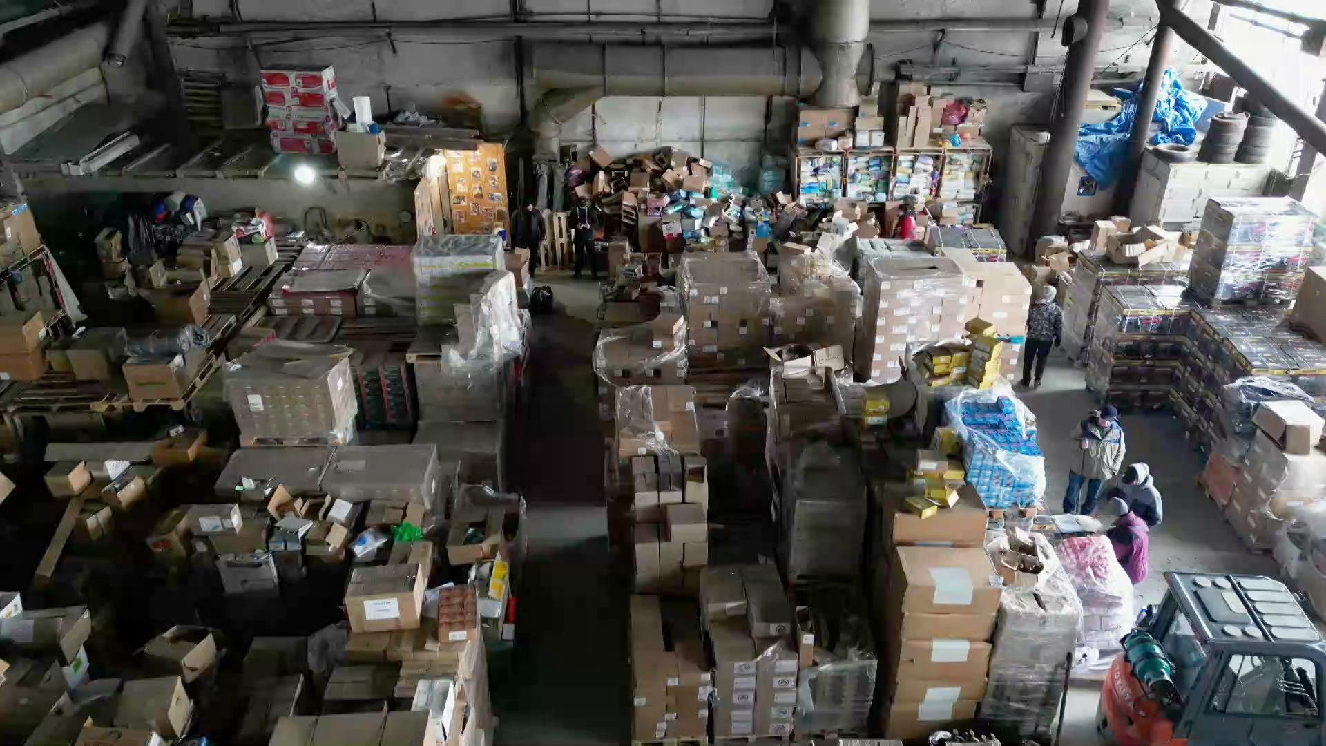 A still image from a video shows an overhead view of a warehouse in Ukraine with several individuals standing among stacks of crates and boxes, and a forklift.