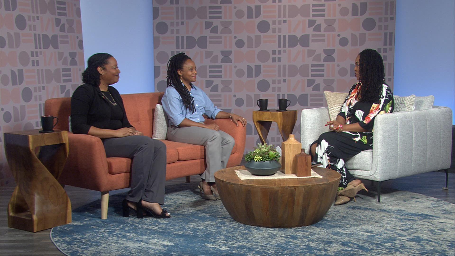 Three women sit in a studio lounge setting, two on an orange sofa and one on a light gray chair, talking across a wooden coffee table.
