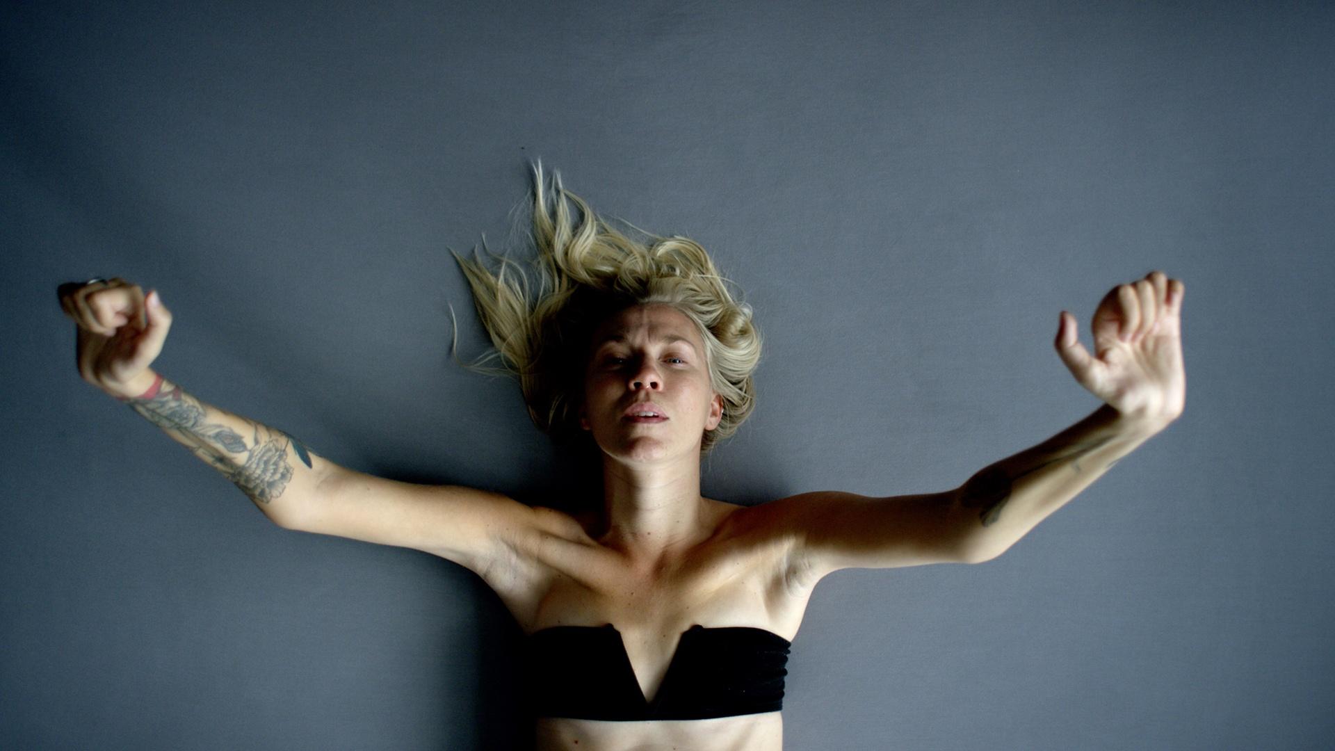 A woman lies on a gray background with her arms outstretched, reaching up towards the camera above her