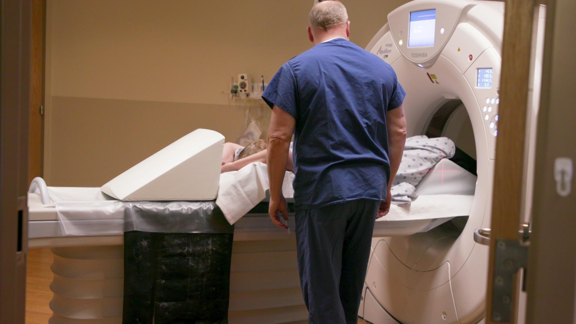 A still image shows a worker in blue scrubs standing over a patient lying down in an MRI machine.
