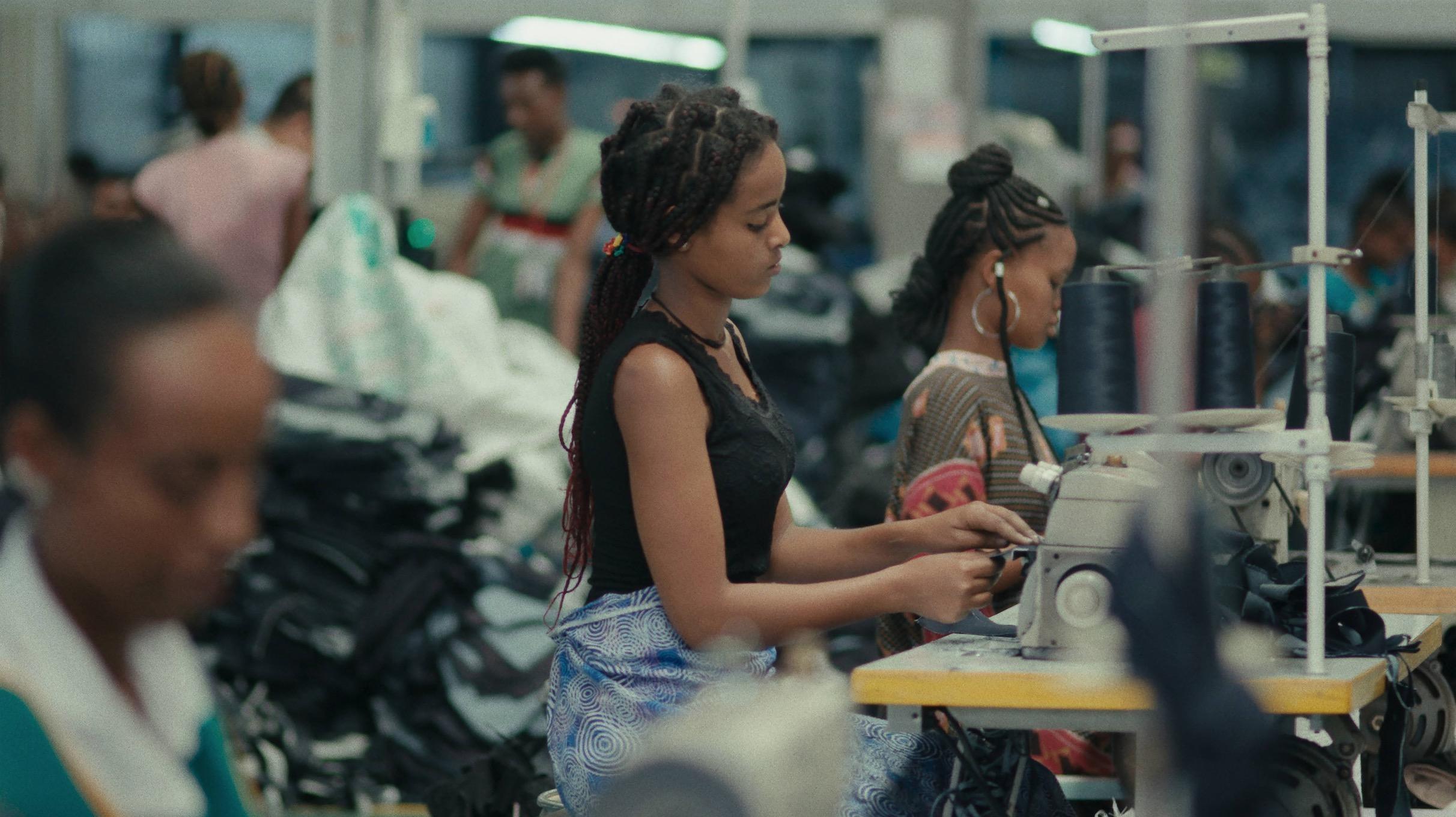 Black women sit at sewing tables in a factory warehouse. 